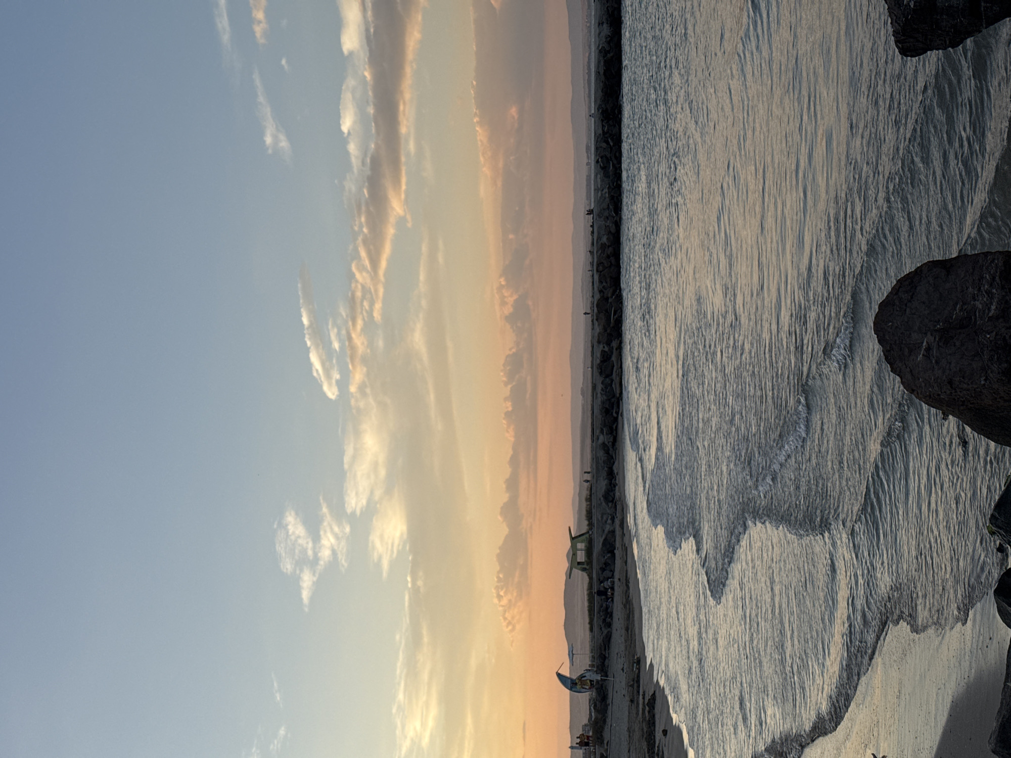 The image depicts a serene sunset scene at a beach in Pomorie, Bulgaria. The sky is painted with hues of orange, pink, and pale blue, with scattered clouds. The calm sea gently laps at the sandy shore, with small waves creating patterns on the water's surface. In the distance, a rocky breakwater extends into the sea, with several people visible on it. Behind the breakwater, a low-lying landscape with rolling hills can be seen. On the beach, there are a few beach umbrellas and a small structure that appears to be a lifeguard station or a small kiosk. A few individuals are present on the beach, some near the water's edge and others further back. The overall atmosphere is peaceful and tranquil, characteristic of an evening at the seaside.
