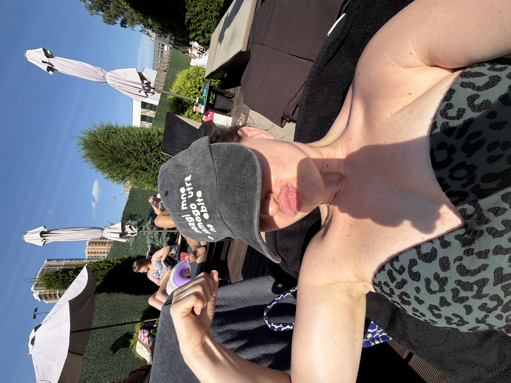 A woman is sunbathing by a pool, wearing a dark green leopard-print bikini top and a black baseball cap that reads "nogi mna samogo utra ne ebite". She is leaning back on a lounge chair, with her arm flexed and her lips pursed. The background shows other people sunbathing, lounge chairs, umbrellas, and palm trees. The setting appears to be a hotel or resort pool area. The sky is clear and blue, indicating a sunny day. The woman is the main subject of the image, and her pose and expression suggest a playful and confident mood. There is a shadow cast on her face and chest from the sun.