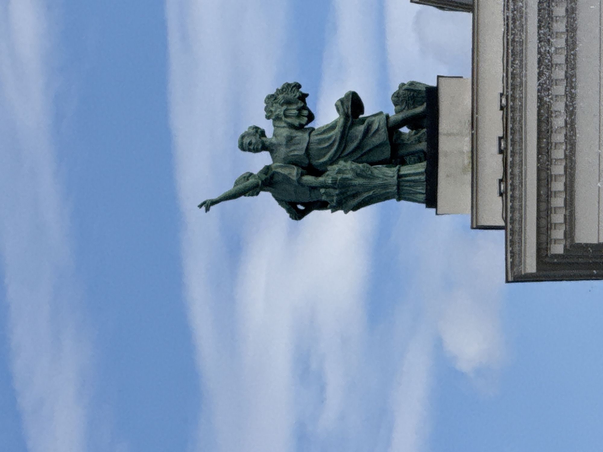 The image shows a close-up, low-angle shot of a bronze sculpture of several figures, likely part of a larger monument, against a bright blue sky with wispy clouds. The sculpture appears to depict a group of women in flowing robes, with one figure in the foreground reaching out her arm. The figures are rendered in a classical style. To the right of the sculpture, a portion of a grey, columned building with decorative elements is visible. The lighting suggests it is daytime, possibly midday, given the bright sky and shadows. The overall scene evokes a sense of grandeur and historical significance. While the exact location is not specified in the image, the prompt provides context that it is Moscow, Russia. No text is visible in the image.