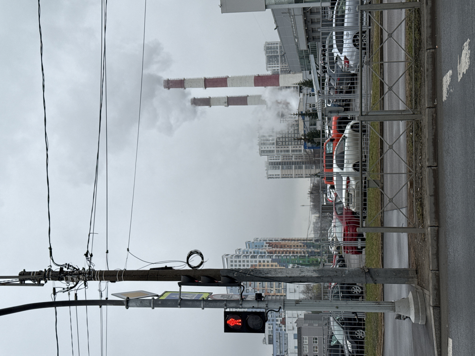 The image depicts an urban landscape under an overcast sky. In the foreground, a utility pole is laden with electrical wires and traffic signals, one of which is displaying a red arrow, indicating "no turn on red". The side of a road is visible, with parked cars behind a metal barrier. In the background, industrial chimneys are emitting smoke, contrasting with the surrounding residential and commercial buildings. It is most likely in the city of Saint Petersburg, Russia, where chimneys like that are common.