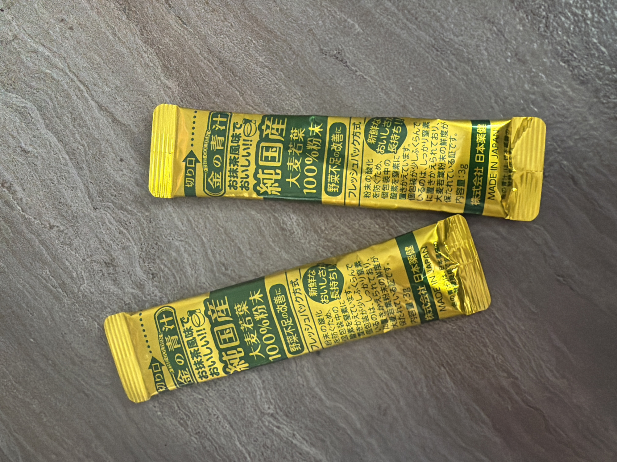 The image shows two yellow and green foil packets of what appears to be a dietary supplement, likely powdered greens. The packets are oriented vertically and are lying on a textured, grey surface that resembles a stone countertop or floor.
The text on the packets is in Japanese and indicates the product is "Golden Aojiru" (金 の 青 汁), described as having a matcha (green tea) flavor and being delicious. It also mentions "domestic production" (純国産), "barley grass" (大麦若葉), and "100% powder" (100% 粉末). There are claims about improving vegetable deficiency and using a "fresh pack method" (フレッシュパック方式) to maintain freshness and nutrients. The net weight is listed as 3g (内容量: 3g). The manufacturer is "Nihon Yakuken Co., Ltd." (株式会社 日本薬健) and the product is "MADE IN JAPAN".
There is no information in the image that can place this scene in Yuzhno-Sakhalinsk, Russia. The products are explicitly labeled as "MADE IN JAPAN".