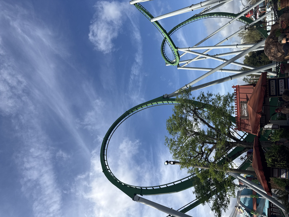 The image displays a section of The Incredible Hulk Coaster, located at Universal's Islands of Adventure in Orlando, Florida, United States of America. The prominent green roller coaster track features a large vertical loop on the left and a series of twists and smaller loops on the right, all supported by numerous inclined gray steel columns. The sky is clear blue with scattered wispy white clouds. In the mid-ground, a large deciduous tree with green foliage stands beneath the primary loop. Below the coaster structure, several theme park elements are visible: a small building with a red roof and a sign reading "NAME ART," two reddish-brown market umbrellas, and portions of other structures, including a teal-domed element on the far left. A tall black pole equipped with two white floodlights is positioned near the large tree. Several indistinct human figures are visible beneath the umbrellas and in the lower foreground, suggesting pedestrian activity within the park. One person's head and shoulder are visible in the bottom right, wearing sunglasses. The scene is brightly illuminated by daylight.