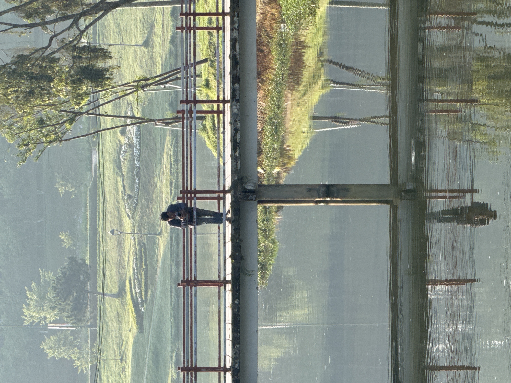 A man is sitting on a bridge, looking at something in his hands. The bridge has a metal railing on both sides. On the left side of the bridge, there is a grassy area with trees. On the right side of the bridge, there is water, possibly a lake. The reflection of the bridge and the landscape is visible in the water. The city and location is Da Lat, Vietnam.