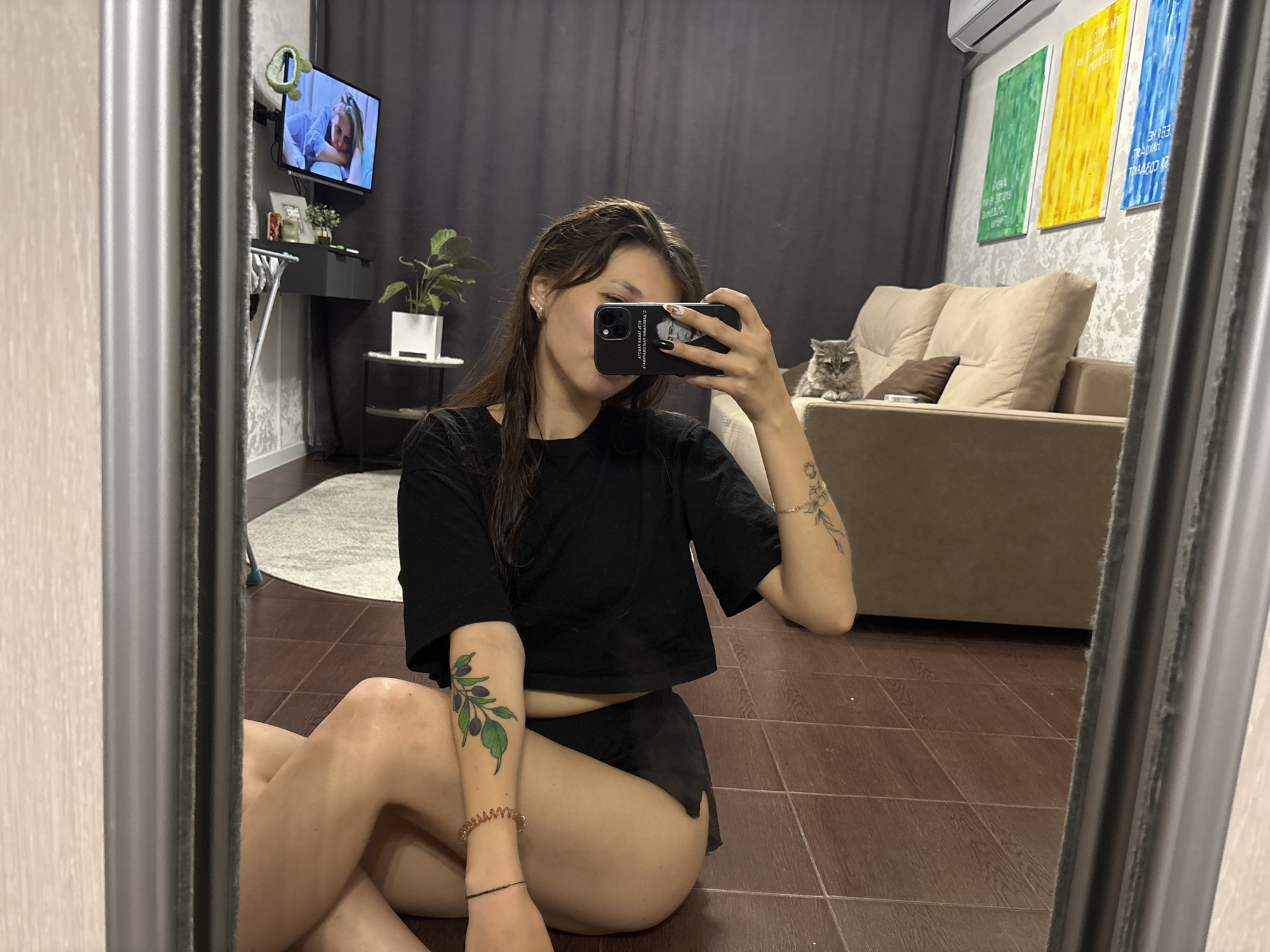 A young woman with dark brown hair is taking a mirror selfie while sitting on the floor. She is wearing a black crop top and black shorts. She has a tattoo of olives on her left arm and a tattoo of flowers on her right arm. She is holding a black smartphone in her right hand. In the background, there is a gray couch with pillows, a cat, a television, a plant, and three colorful paintings on the wall. The room has wooden floors and a gray curtain. This scene is located in a residential apartment in Novokuznetsk, Russia.