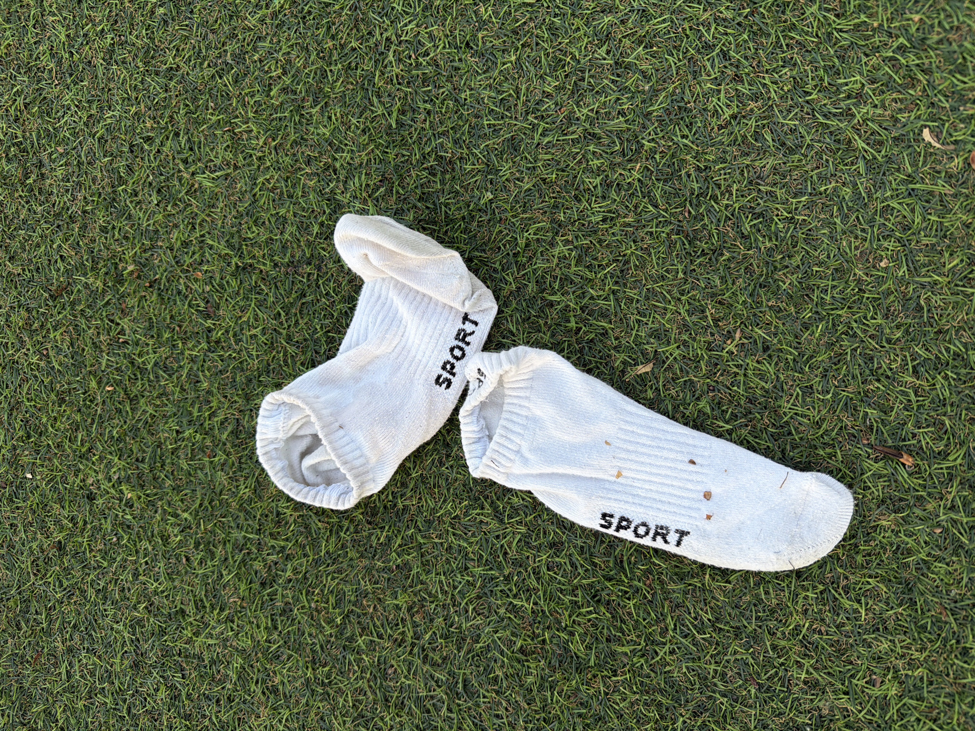Two white sport socks lie on green grass. The word "SPORT" is printed on each sock in black lettering. The socks are slightly soiled with small bits of debris. The setting appears to be outdoors on a grassy surface, possibly a lawn or field. There is no indication of the city of Moscow, Russia, or any people in the image.