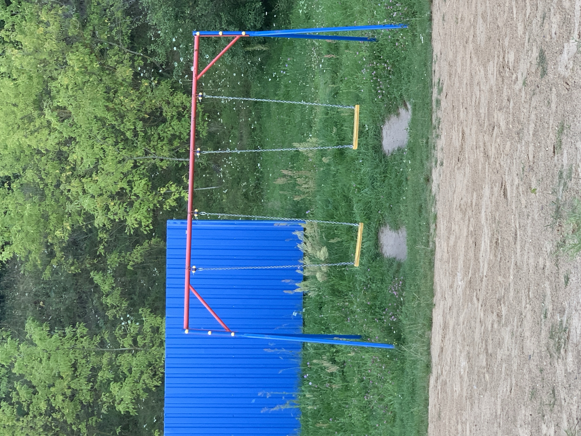 A metal swing set with a red top bar and blue support legs stands in a grassy area. Two wooden swings hang from chains attached to the top bar. A bright blue corrugated metal fence is visible behind the swing set, with a dense green forest in the background. The ground in front of the swing set is a sandy, dirt surface. The scene appears to be a playground or park area. This location is identified as Kapyĺ, Belarus.