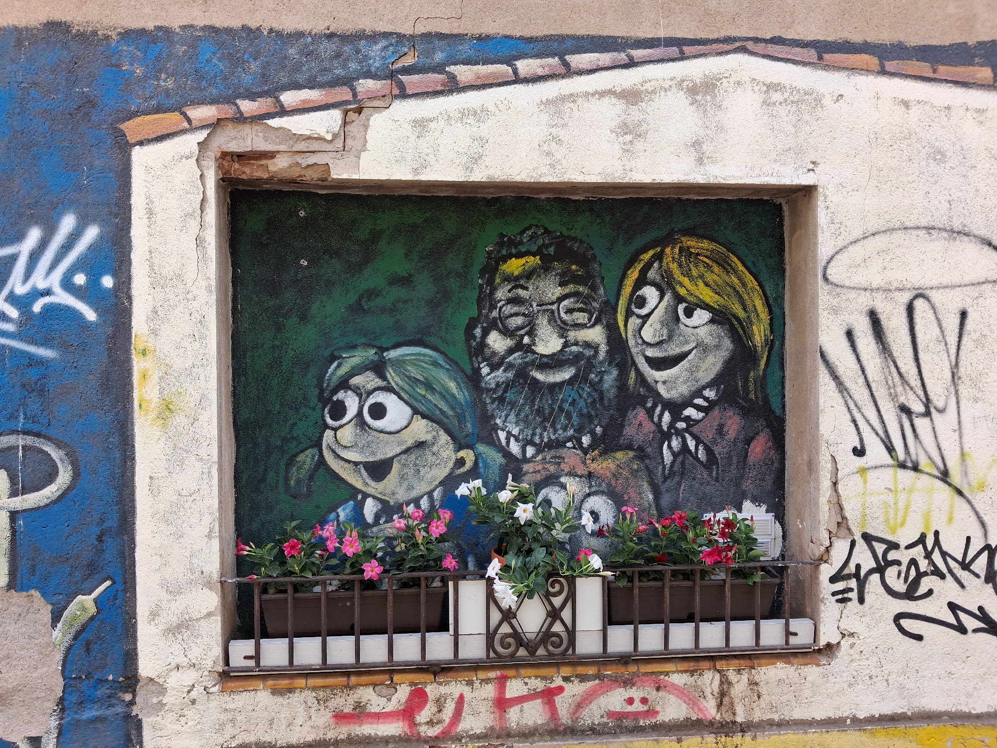 The image shows a mural on the exterior of a building in the city of Salt, Spain. The mural depicts three cartoon-like figures with large eyes and wide smiles. The figures are painted against a dark green background. In front of the mural, there is a window with two brown planters filled with pink and white flowers. A metal railing separates the planters from the bottom of the window. The building's facade is weathered and shows signs of damage, with cracks in the plaster and peeling paint. Graffiti in various colors covers parts of the exterior wall, including the area around the window. The overall scene suggests a street art display on a public building in an urban environment.