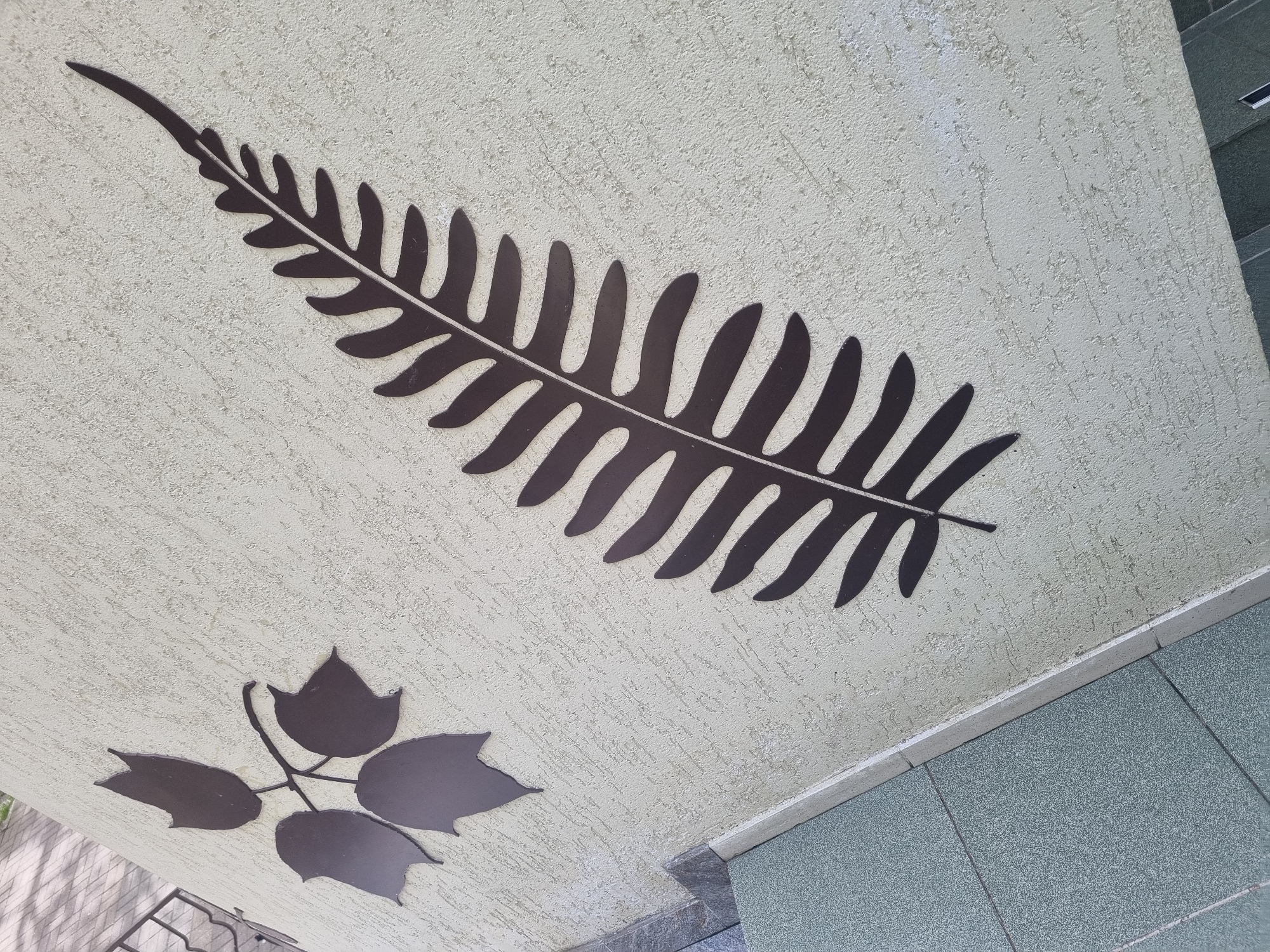 A high-angle, full-shot photograph shows a section of an exterior wall with decorations. The wall is painted in a light beige color with a textured surface. A large, dark brown fern leaf design is prominently displayed, oriented diagonally. Below and to the left of the fern leaf is a smaller design of a maple leaf arrangement with four leaves. The bottom right corner of the image shows a transition to a tiled surface of gray, suggesting a sidewalk or patio. The location is Sochi, Russia.