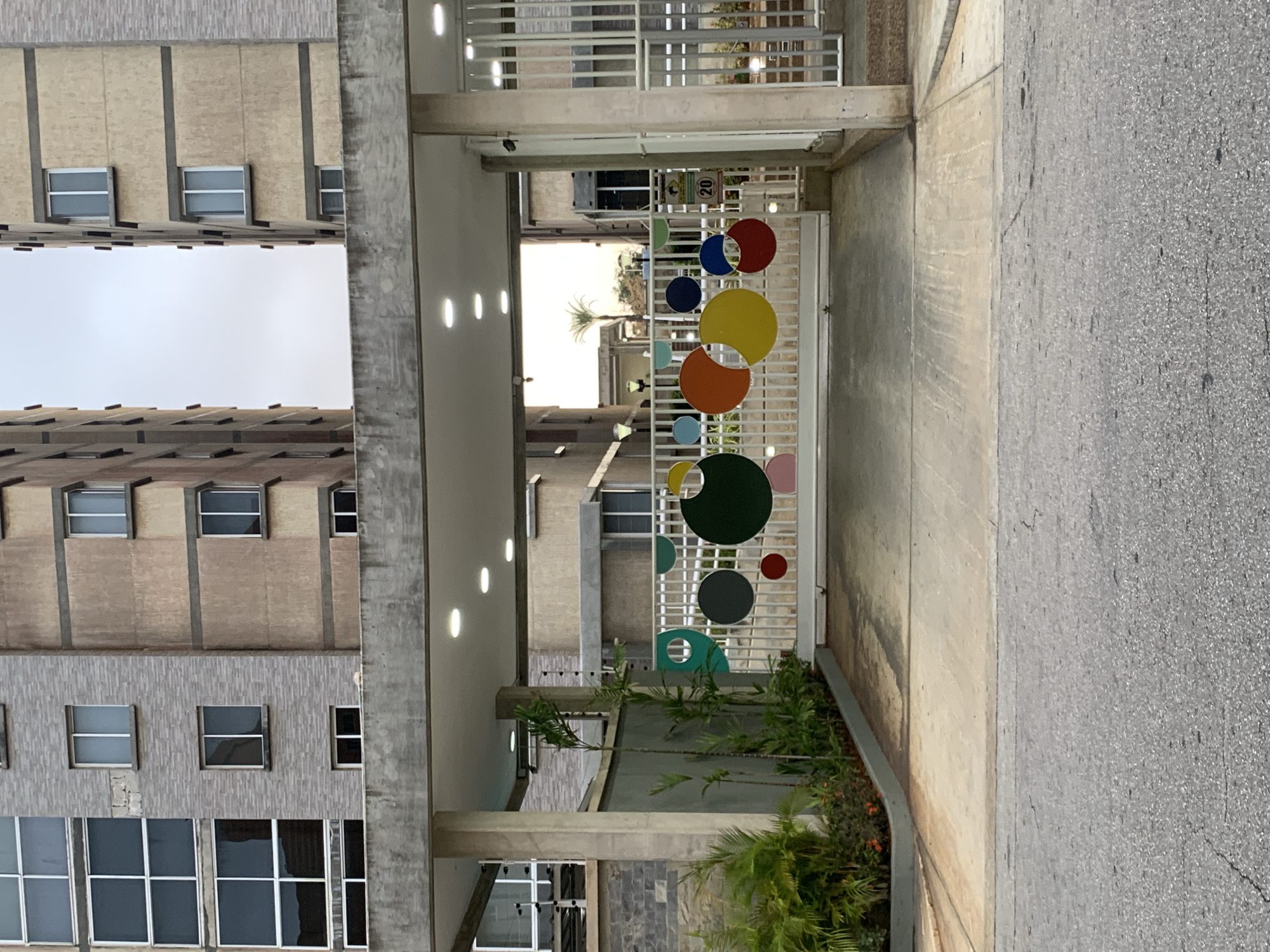This media file, oriented 90 degrees clockwise, captures the entrance to a building complex in Pampatar, Venezuela, under bright daylight.
The scene is dominated by two multi-story buildings flanking a central covered entrance. The buildings, visible on the left side of the frame (top in the rotated image), feature a functional architectural style with facades composed of both light brown and gray textured sections. They have numerous rectangular windows, often with white frames, arranged in repetitive patterns. A bright, clear sky is visible in the open space between these structures.
At the center of the image, a white metal gate acts as a vibrant focal point. It is decorated with an array of colorful circular cutouts of various sizes, including red, blue, yellow, orange, dark green, light green, light blue, and pink, lending a playful and modern touch to the entrance. Behind the gate, a covered concrete walkway leads into the complex, illuminated by several recessed lights on its ceiling. Through the gate, a glimpse of distant buildings and a tropical palm tree can be seen, hinting at the surrounding environment. A small yellow diamond-shaped sign with the number "20" in black text is visible near the gate, likely indicating a speed limit or similar regulation.
To the right of the gate (bottom in the rotated image), a grey, paved asphalt road or pathway runs parallel to the building. A concrete curb separates the road from a small, manicured garden area filled with lush green plants and shrubs, providing a natural contrast to the concrete and asphalt.
The scene appears quiet and devoid of people, suggesting a calm moment during the daytime under clear weather conditions. The utilitarian architecture combined with the artistic, colorful gate presents an interesting visual contrast typical of a modern urban residential or commercial development in a tropical setting.