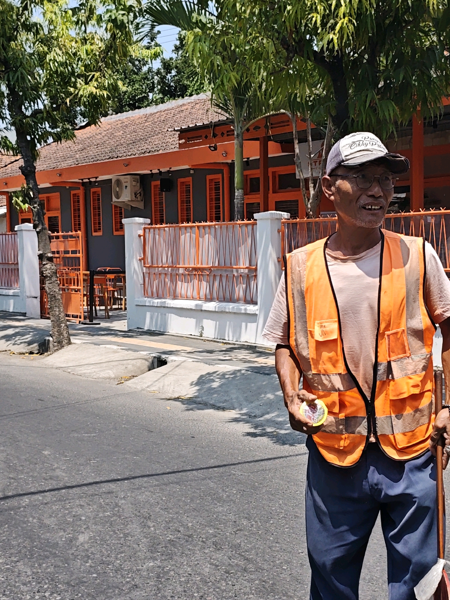 In the city of Kediri, Indonesia, a male street sweeper stands on the side of a paved road, wearing a light-colored t-shirt, dark blue pants, a reflective orange vest, and a cap. He is holding a small, round yellow object in his right hand and a broom in his left. The background features a building with an orange facade and roof, a white fence with orange metal bars, and trees. The scene appears to be captured during daylight.