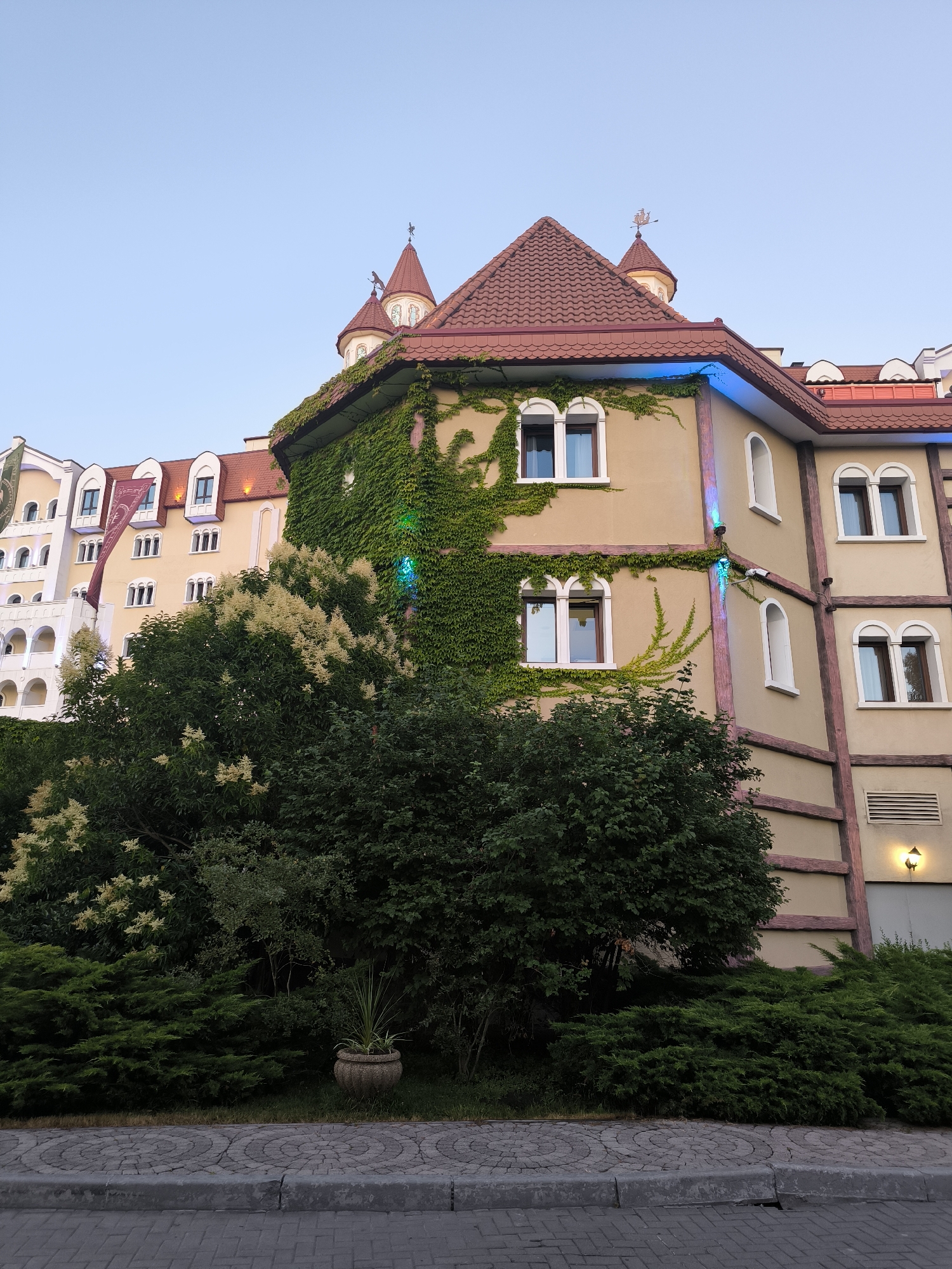 The image depicts a building with a distinctive architectural style, featuring arched windows, a red-tiled roof, and a turret topped with a weather vane. The building is adorned with climbing ivy, creating a lush green facade. In the foreground, there are various trees and bushes, and a cobblestone pavement. The sky is clear and blue, suggesting it is daytime. The building appears to be a hotel or resort, possibly part of the Sirius federal territory in Russia, given its elaborate design and landscaping. There are no people visible in the image. The scene is well-lit, with natural light illuminating the building and vegetation.