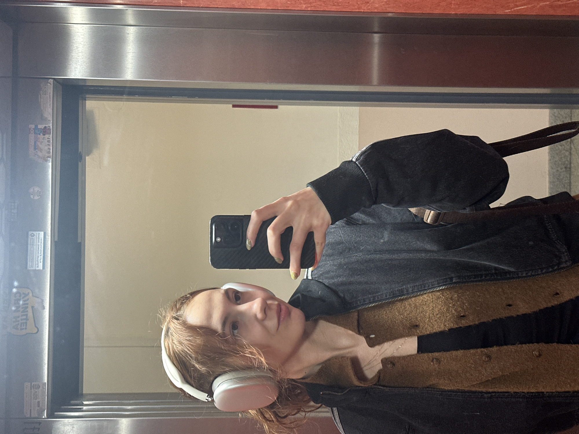 A person is taking a selfie in what appears to be an elevator. The person has light skin, reddish-brown hair, and is wearing white headphones, a brown cardigan, a black top, and a dark denim jacket. The person is holding a smartphone with a black case and light green nail polish. The elevator has silver walls and a red top, with stickers present on the left side. The image is rotated, giving a vertical perspective. The environment suggests an indoor location. There is no specific information in the image to determine if it is Novi Sad, Serbia.