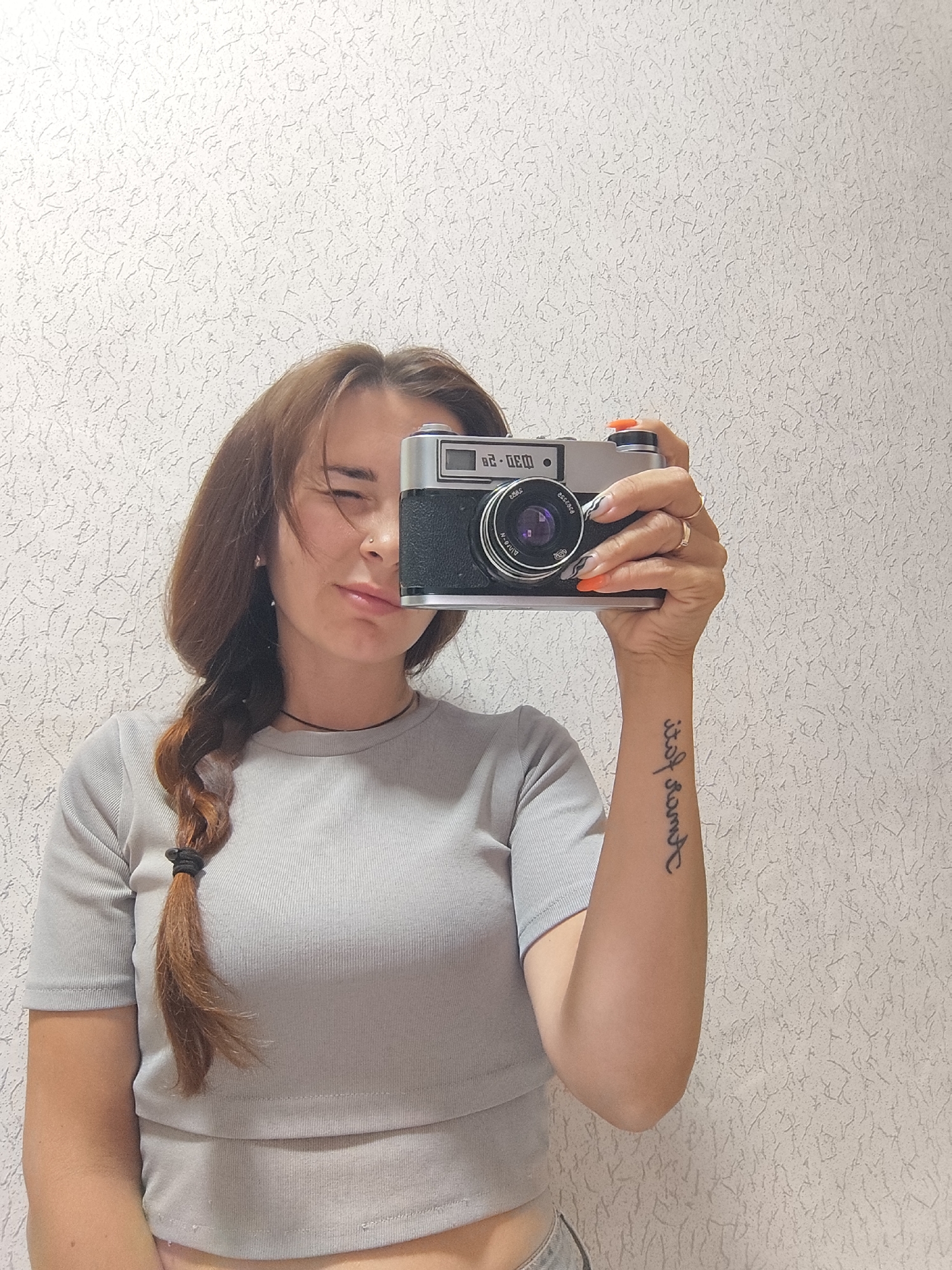 A woman is holding a vintage film camera up to her face, partially obscuring it. She has light brown hair styled in a braid that falls over her shoulder. Her right hand is holding the camera, and her left hand is visible supporting it. She is wearing a light gray, ribbed crop top. Her left eye is partially closed, and she has a playful expression. She is also wearing a ring on her right ring finger and has bright orange nail polish on her fingernails. A tattoo is visible on her left forearm. The background is a textured, light-colored wall. The scene appears to be indoors, possibly in a home in Фоки, Russia. There is no indication of the time of day or weather conditions. The visible text on the camera is "030-20".