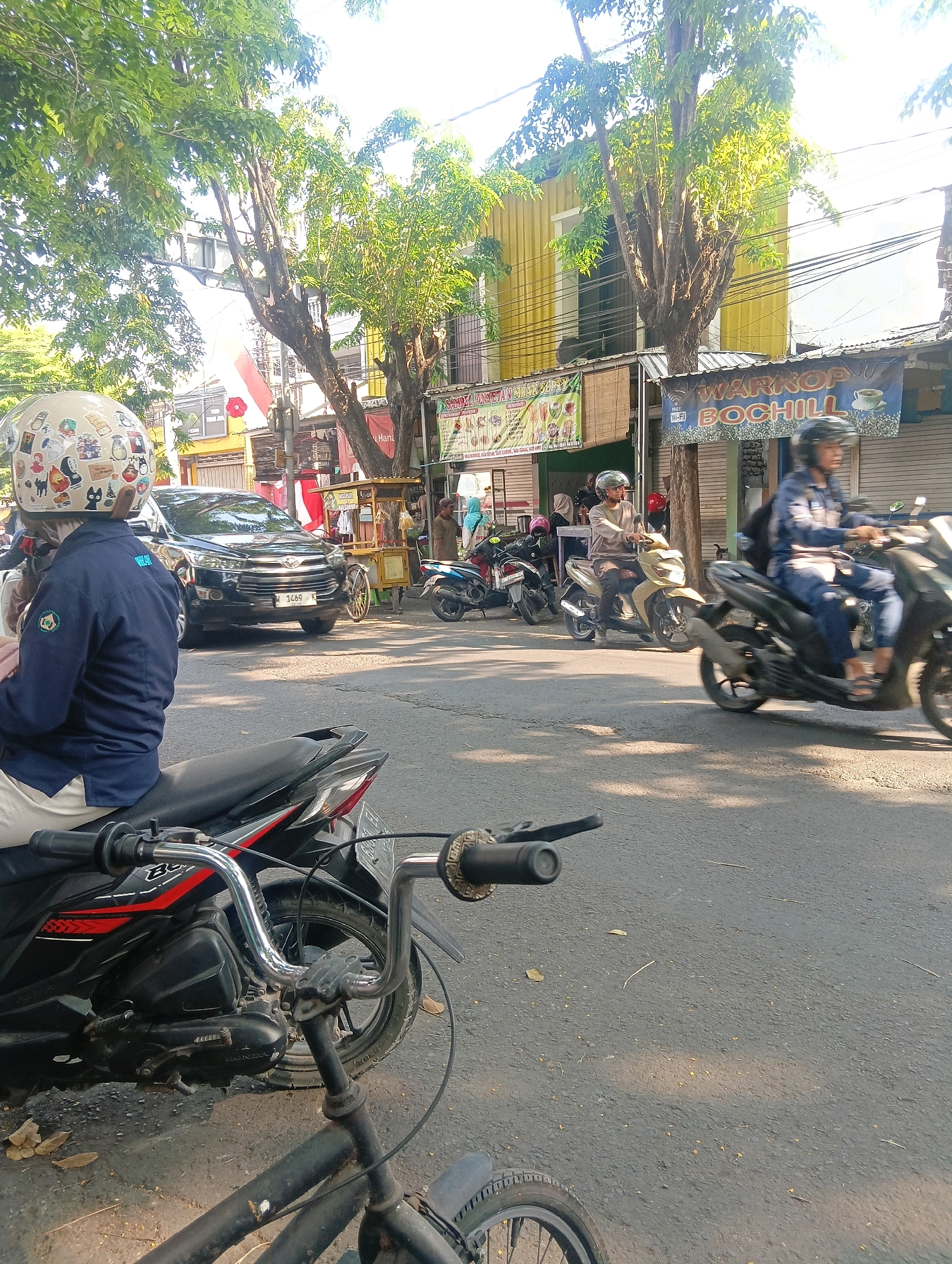 This image depicts a street scene in Gapurosukolilo, Indonesia. The foreground features the handlebars and front wheel of a bicycle, with a black motorcycle positioned behind it, its engine and rear tire partially visible. In the mid-ground, several people are on motorcycles, with one person in the foreground on a motorcycle wearing a blue jacket and a helmet adorned with stickers. To the right, a person in a blue shirt and helmet is riding a motorcycle. A black car with license plate "1469" is stopped in traffic. In the background, there are several buildings, including one with a yellow facade and another with a banner that reads "WARNOP BOCHILL" and indicates "WI-FI." Trees line the street, providing shade. The overall scene suggests a busy street with vehicular traffic and commercial establishments.