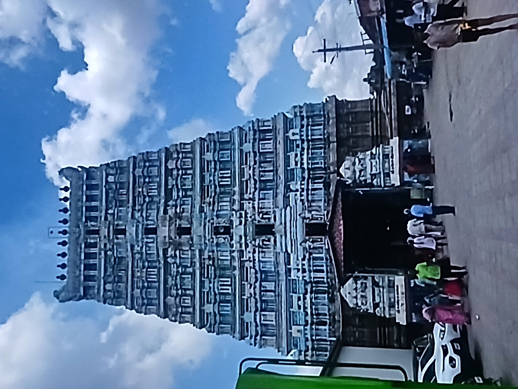 The image depicts a tall, ornate temple gopuram (tower) in Bengaluru, India, under a blue sky with white clouds. The tower is multi-tiered with numerous architectural details and is light gray in color. The base of the tower transitions into the temple entrance where people are milling around. The scene includes pedestrians, including people wearing colorful clothing, walking on a paved street adjacent to the temple. A white car and a green vehicle are visible in the foreground, partially obscuring the temple entrance. A power pole is visible near the top edge of the image.