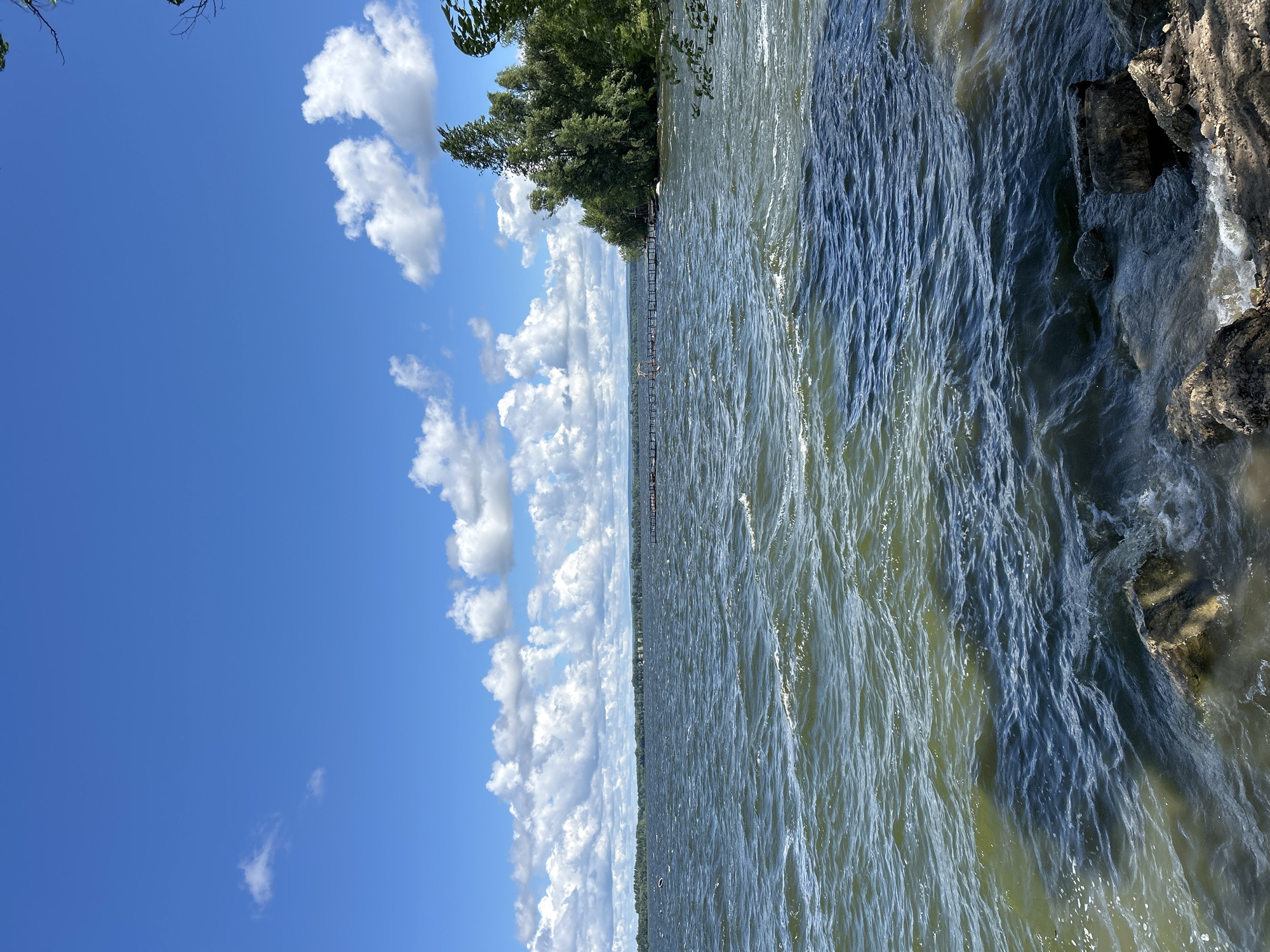The image depicts a view of a wide body of water on a sunny day, under a bright blue sky with scattered white cumulus clouds. The water is choppy, with visible waves. In the foreground, there are rocks along the shore. Across the water, a long wooden pier or walkway extends from the treeline on the right side of the image. The treeline appears dense and green. The far shore is visible in the distance, a hazy line of trees and vegetation against the horizon. The scene suggests a natural outdoor setting, likely a lake or a wide river. There are no people or animals visible in the image. The location is not identifiable as being in the city of Междуреченск, Russia.