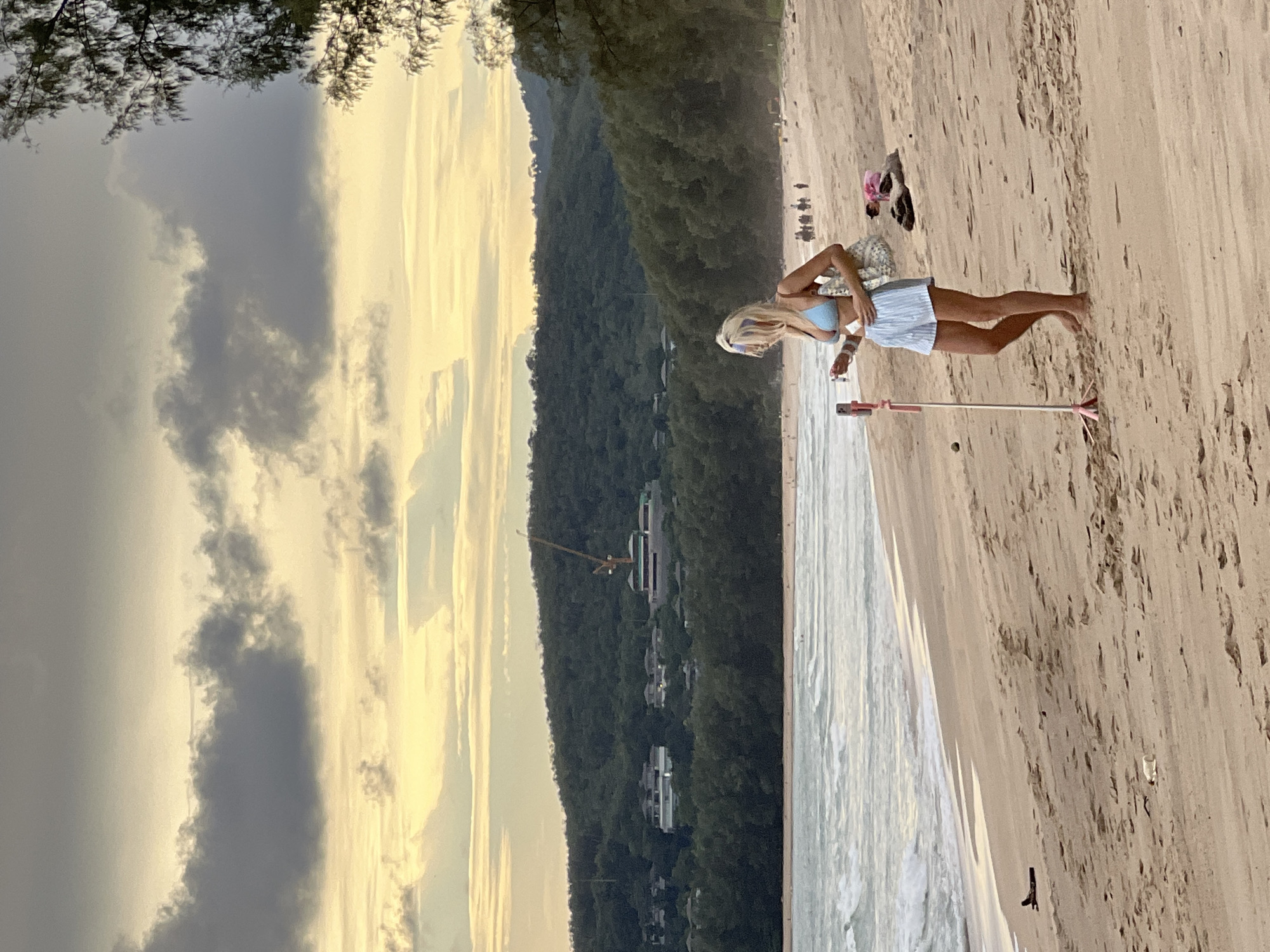 A blonde woman in a light blue bikini top and striped shorts stands on a sandy beach, holding a phone attached to a selfie stick. She is looking down at her phone, which is positioned on the tripod. Behind her, the ocean waves roll onto the shore, and further back, a lush, green hillside rises, dotted with buildings and a construction crane. The sky above is filled with dramatic clouds tinged with the warm hues of sunset. Other people are visible in the distance on the beach and in the water.