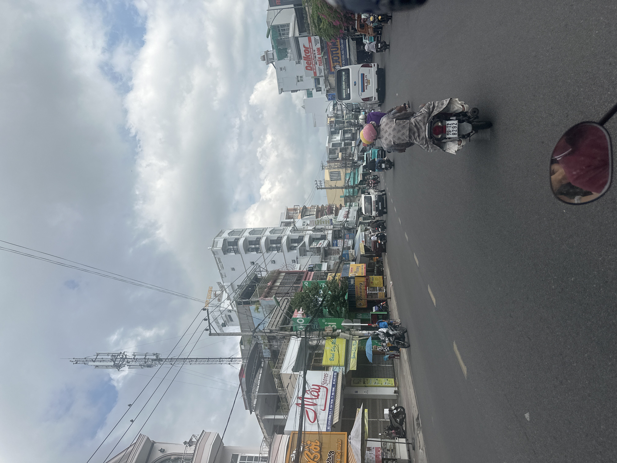 The image is an eye-level, slightly tilted view of a street scene in Nha Trang Ward, Vietnam. The road is asphalt, marked with intermittent white lines. A line of motorbikes fills the street; many are moving. On one motorbike, a rider and passenger are covered in a grey fabric, the license plate reads "79-T2 8292". The passenger wears a multicolored helmet.
Buildings line the left side of the street, densely packed together. Many have shop signs, with Vietnamese script. The sign for "May Stores" is visible, advertising the address "1218 Đường 2/4 - P. Vạn Thắng - TP. Nha Trang". Other signs read "Delkor" and "JOTUN". A network of electrical wires stretches across the sky, intersecting with a communication tower. The sky is partly cloudy. A motorcycle side mirror is visible in the right edge of the image, reflecting the person who took the photograph.