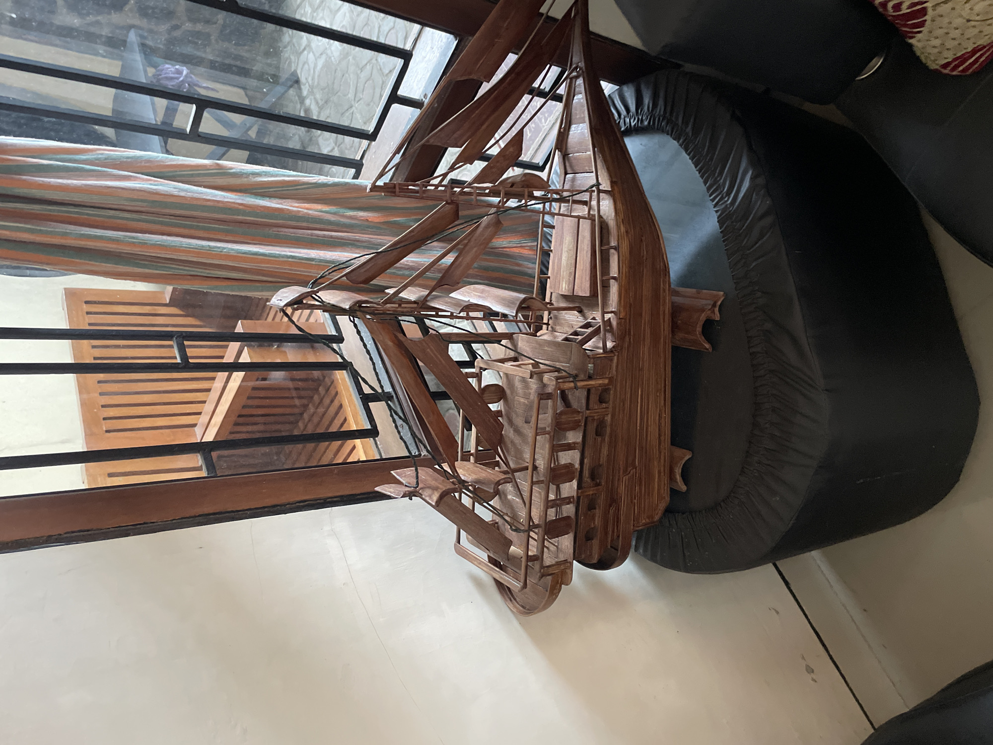 A wooden model of a sailing ship is displayed on a black ottoman, positioned in front of a window with vertical blinds and a wooden slatted chair. The ship is intricately detailed with masts, sails, and a deck. The scene appears to be an indoor setting, possibly a home or shop in Pekalongan, Indonesia, judging by the presence of traditional Indonesian furniture and decor. The lighting is natural, coming from the window, casting shadows on the ship and the surrounding objects.
