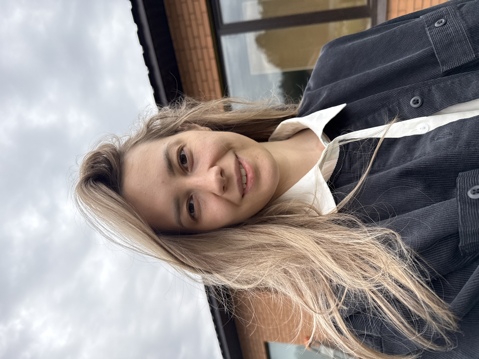 A woman with long, blonde hair is smiling at the camera. She is wearing a white collared shirt underneath a dark blue, corduroy jacket. The background includes a cloudy sky and a brick building with large windows. The exact location within the city of Chaikovsky, Russia, cannot be determined from the image alone.