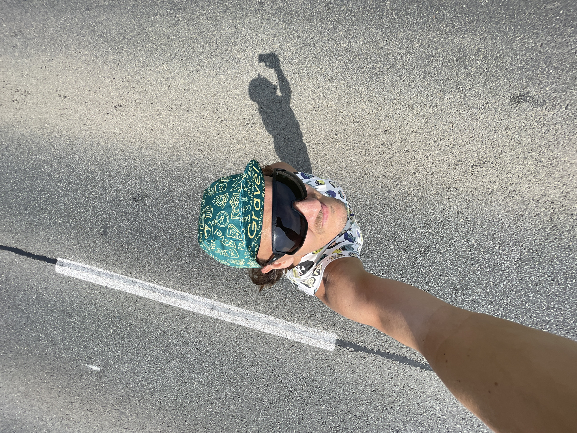 A high-angle selfie taken outdoors on asphalt, possibly in the city of Perevoznoe, Russia, shows a man wearing sunglasses and a green cap with a patterned design. He is wearing a white t-shirt with a graphic print. The man's arm is visible, extended upwards to capture the shot, and his shadow is cast on the ground to his right. A white line runs diagonally across the asphalt, and a black line appears to be on the left side of the image. The scene is illuminated by bright sunlight, creating a distinct shadow of the man.