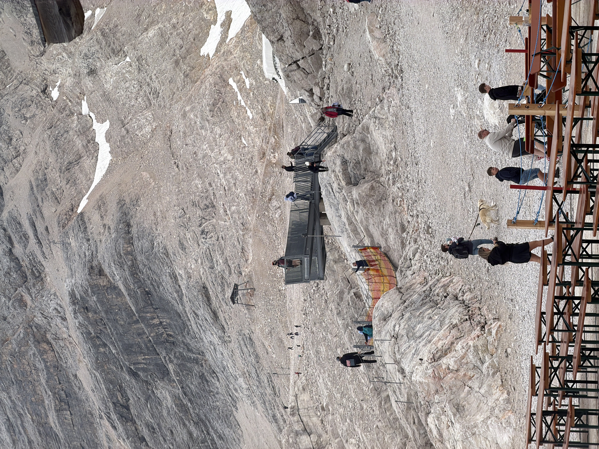 The image depicts a mountainous, rocky terrain with scattered snow patches and sparse vegetation, characteristic of a high-altitude environment near Garmisch-Partenkirchen, Germany. In the foreground, several rows of empty wooden picnic tables and benches are set up on a gravelly surface, suggesting a designated visitor area.
Further up the slope, a metal walkway or viewing platform is visible, with several people standing and walking on it. Some individuals are descending a set of metal stairs from the platform. A safety fence, constructed with orange netting and metal poles, guides visitors along a path.
Numerous people are scattered across the rocky landscape, some walking, others standing and observing. One person is walking a golden retriever on a leash. The general atmosphere is one of tourism and recreation in a natural, mountainous setting. The rugged, layered rock formations dominate the background, indicating a significant elevation.