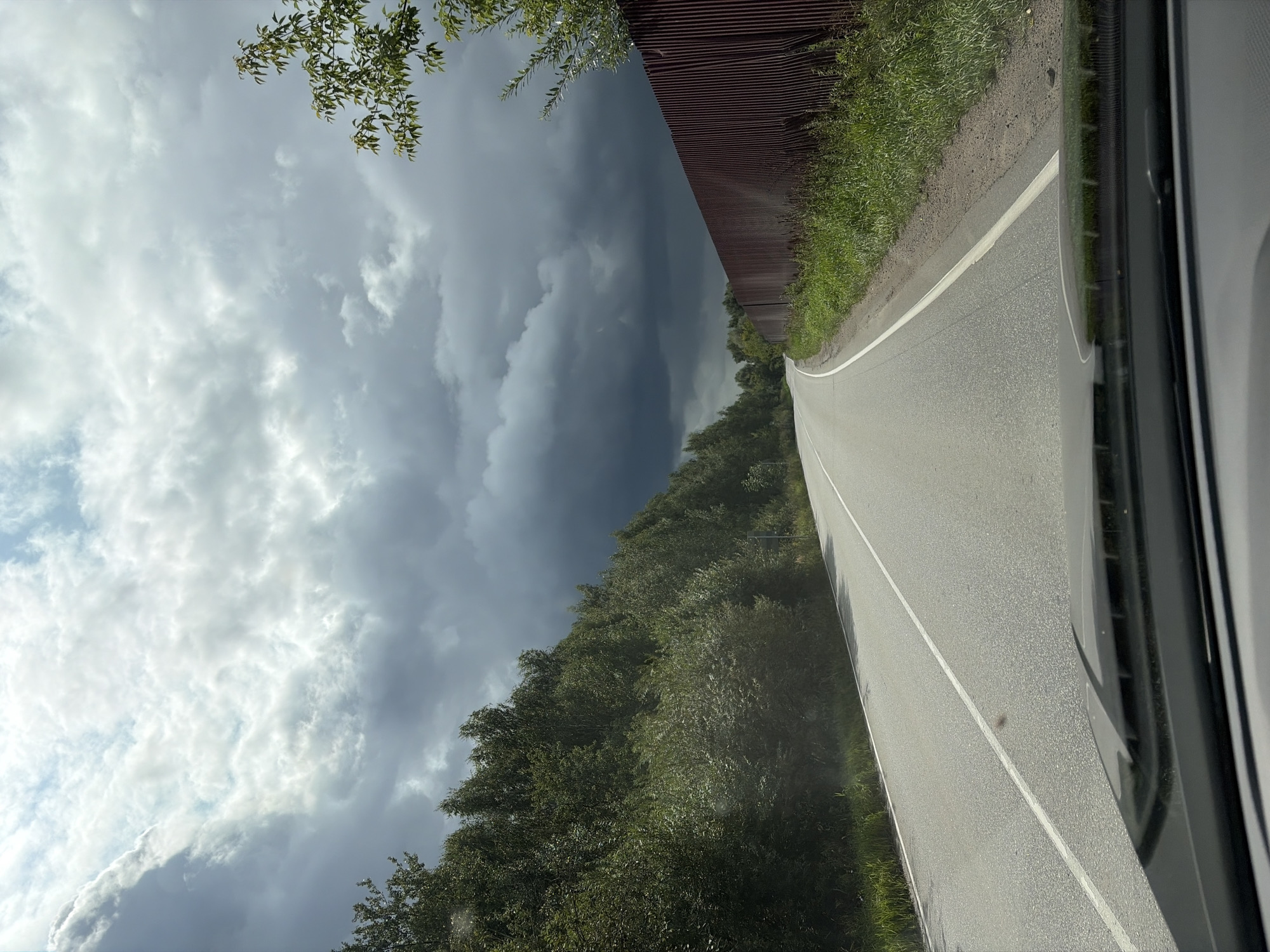 The image shows a view from inside a vehicle traveling on a paved road, likely in the city of Imeni Vorovskogo, Russia. The road curves to the left and is bordered by a dense line of green trees and foliage on the left and a rusty corrugated metal fence on the right. The sky is overcast with heavy, dark storm clouds dominating the upper half of the frame, with patches of lighter clouds and hints of blue sky visible in the distance. The sun is partially obscured by the clouds, casting a diffused light on the scene. The foreground shows the dashboard and windshield of the vehicle.