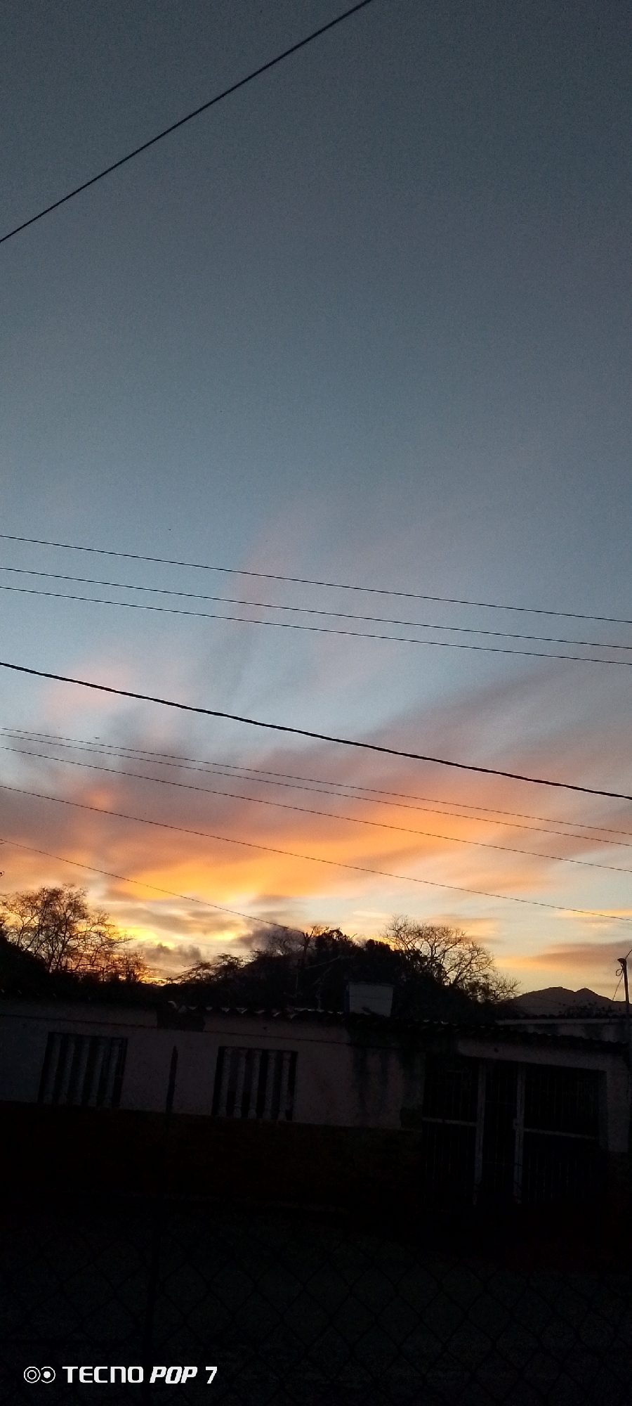 The image shows a sunset over a building in Altagracia, Venezuela. The sky is transitioning from blue to orange, with the orange hues dominant near the horizon. Power lines run across the frame. The building is silhouetted against the bright sky. The foreground is dark, possibly due to being behind a fence or shrubbery. The TECNO POP 7 watermark is visible at the bottom left corner.