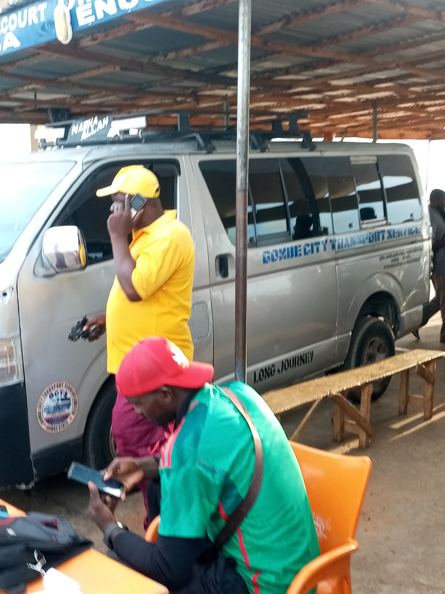 The image depicts a street scene in Gombe City, Nigeria, under a makeshift shelter with a corrugated metal roof supported by wooden beams. In the foreground, a man wearing a red cap and a green and red striped shirt sits in an orange plastic chair, looking at a smartphone. To his left, a black bag lies on an orange table. Behind him, a silver minivan with "GOMBE CITY TRANSPORT SERVICE" and "LONG JOURNEY" written on its side is parked. A man in a yellow polo shirt and yellow cap is standing beside the minivan, speaking on a mobile phone and holding a dark object in his other hand. A sticker with a circular emblem featuring a ship and text is visible on the side of the minivan. The ground appears to be dusty or dirt.