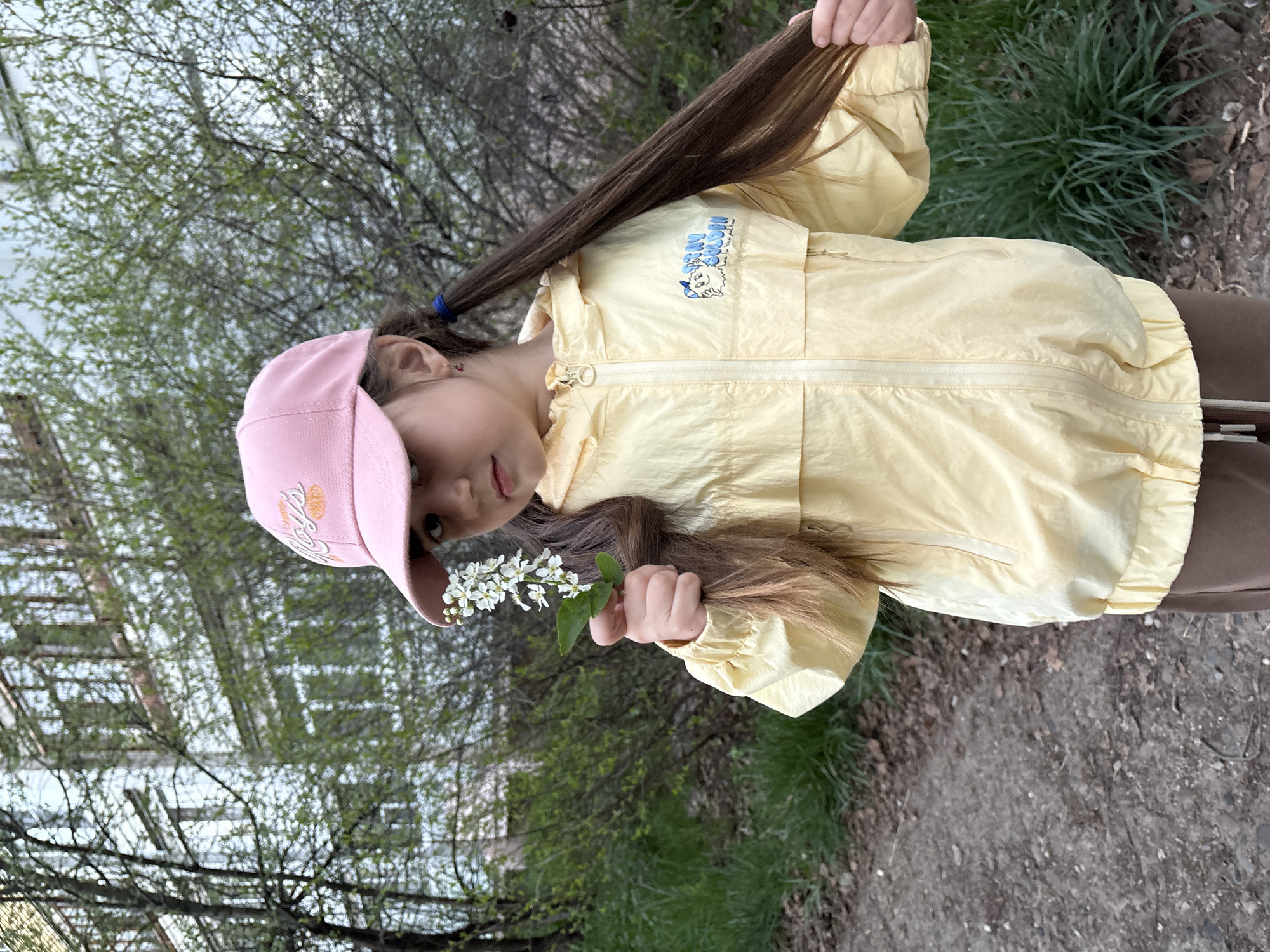 A girl is standing outside in the city of Chaikovsky, Russia. She is holding a sprig of white flowers in her right hand and holding her long, brown hair with her left hand. She is wearing a pink cap, a light yellow jacket, and brown pants. Behind her is a green grassy area with trees and a building. The ground is gray with some dirt.