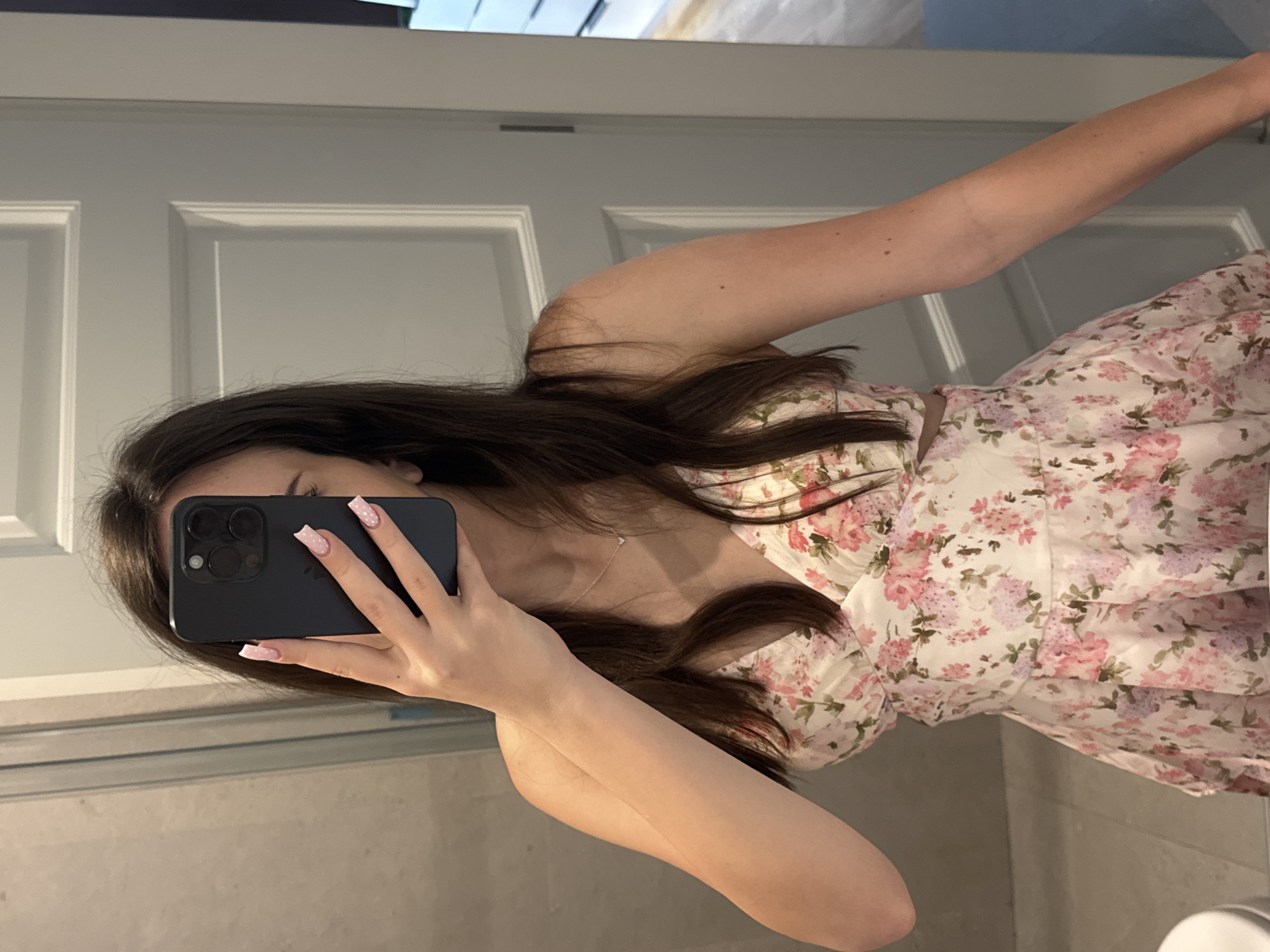 An image taken as a mirror selfie shows a person, likely female, from the chest up, positioned vertically as if looking sideways into a mirror. The person is holding a dark-colored smartphone, possibly an iPhone due to the triple camera configuration, to capture their reflection. The phone obscures the person's face.
The person has long, dark brown hair that cascades over their shoulders and chest. They are wearing a white dress adorned with a vibrant floral pattern featuring pink, purple, and green hues. A delicate silver necklace with a small, clear pendant is visible around their neck. Their fingernails are long, with a square shape, painted light pink with small white polka dots.
The background primarily consists of a white, paneled door, suggesting an indoor setting like a bathroom. The reflective surface of the mirror is prominent, and at the bottom of the frame, there's a hint of a countertop or ledge. The lighting appears to be indoor, and no specific time of day or weather can be determined. There is no visible text in the image.