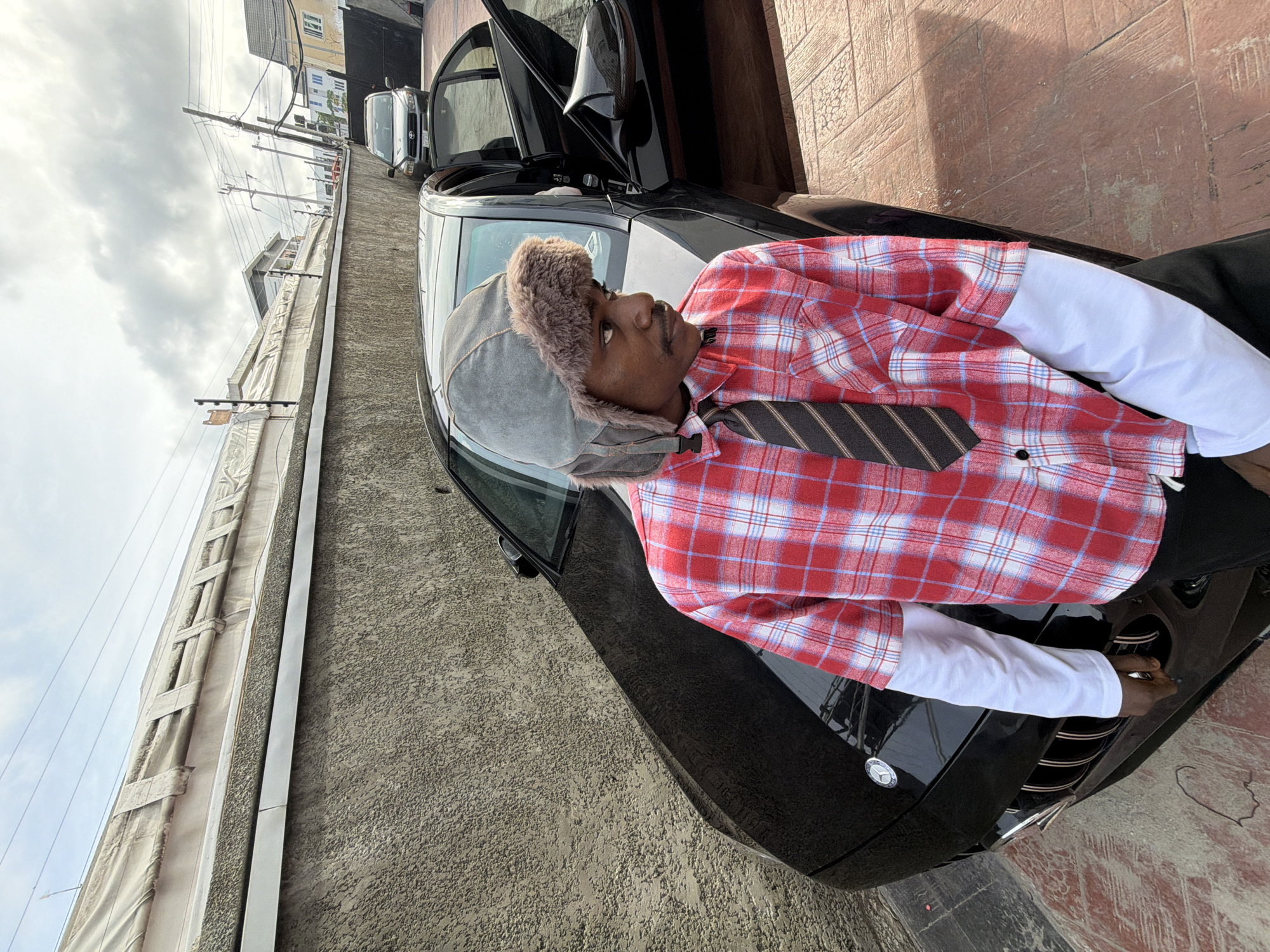 A man wearing a gray fur-lined hat, a red and white plaid shirt over a white undershirt, and a brown striped tie, is posing while leaning against the hood of a black car with its doors open. The setting appears to be an urban area in Igboefon, Nigeria, as suggested by the location. The background includes buildings and power lines. The pavement and the side of a building are visible.