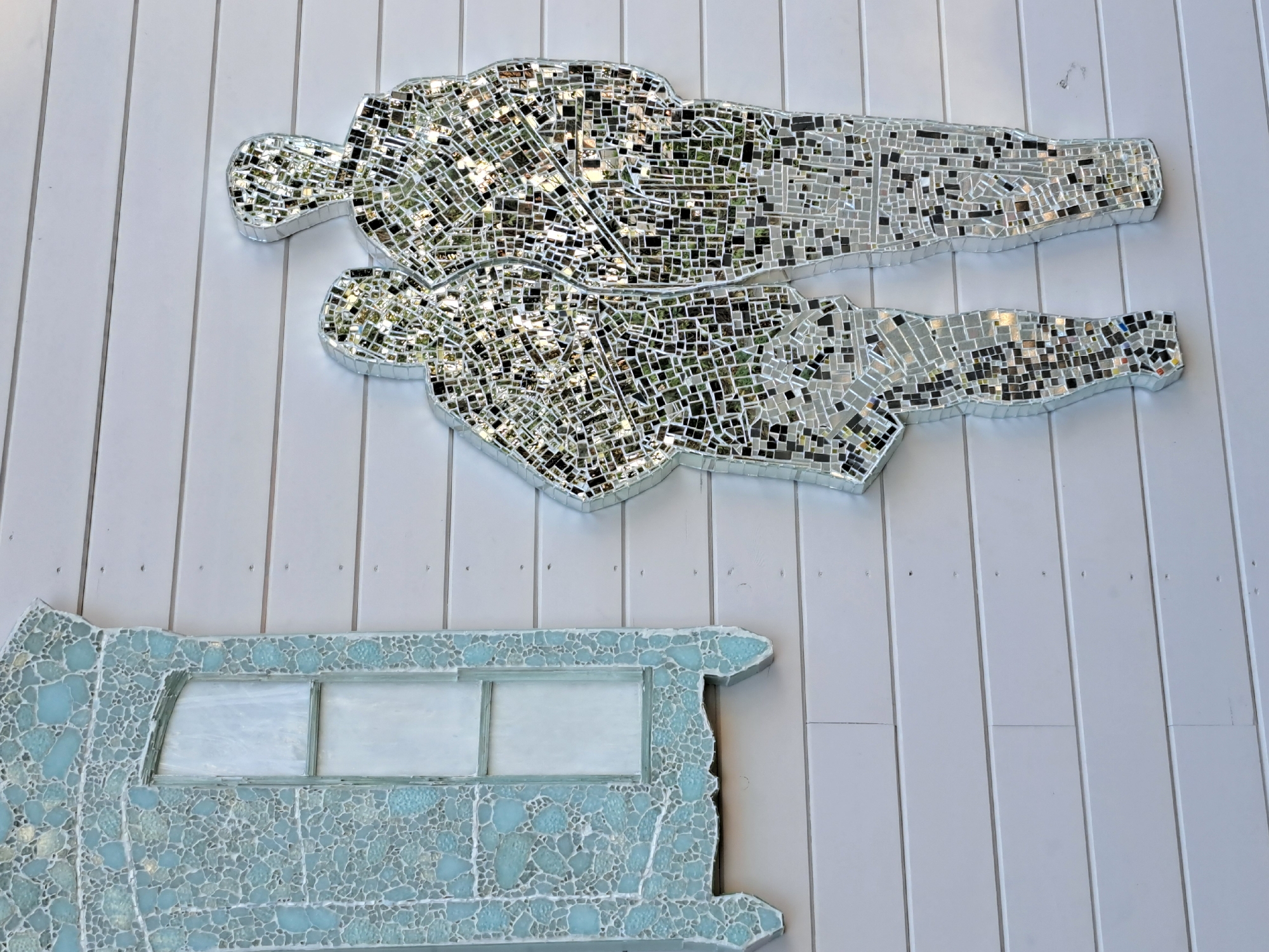 The image shows a wall of white horizontal wooden planks. On the left side of the wall is a mosaic artwork depicting a light blue, ornate window frame with a frosted glass pane. To the right of the window artwork, a mosaic artwork of two human figures, constructed from mirrored tiles, stands side-by-side, appearing to be in conversation. The mirrored tiles reflect the surrounding environment, showing patches of greenery and sky. This artwork is located on the wall of a building in Irkutsk, Russia.