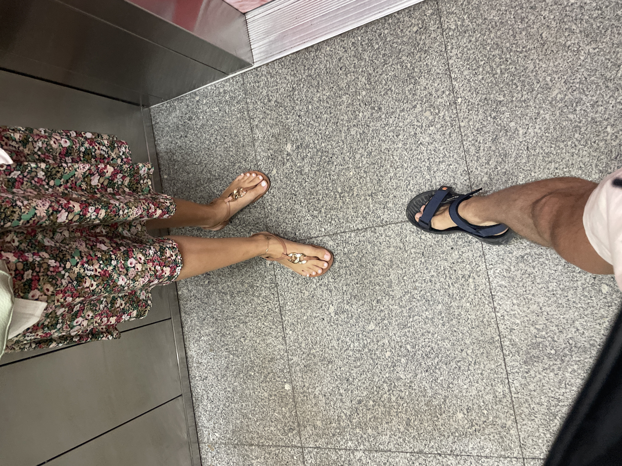A top-down view of two people's legs and feet in an elevator. One person is wearing a floral dress and gold-toned flip-flops. The other person is wearing blue and black sandals, white shorts, and has darker skin. The floor is grey with a speckled pattern. The elevator's interior is made of metal panels. The location is Nha Trang Ward, Vietnam.
