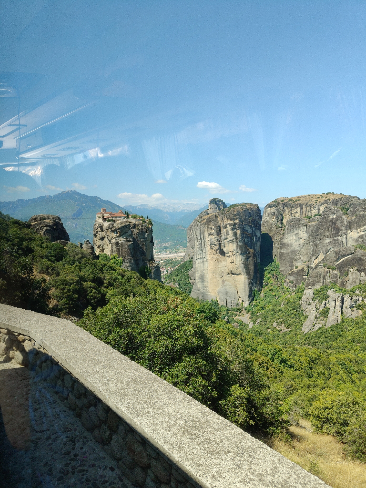 The image depicts a panoramic view of the Meteora monasteries in Kalambaka, Greece. In the foreground, a stone wall with a cobblestone path runs along the bottom of the frame, suggesting a viewpoint or walkway. Beyond the wall, lush green trees and vegetation cover the slopes of the valley. Dominating the mid-ground are the iconic rock pillars of Meteora, with the monastery of Holy Trinity perched atop one of the largest spires. The monastery features a terracotta-tiled roof and a cluster of buildings. Further in the background, several other imposing rock formations rise from the landscape, some with hints of vegetation on their steep sides. The horizon is marked by rolling mountains under a clear, bright blue sky with a few wispy clouds. The scene is bathed in natural sunlight, indicating it is daytime. There are no people or animals visible in the image.