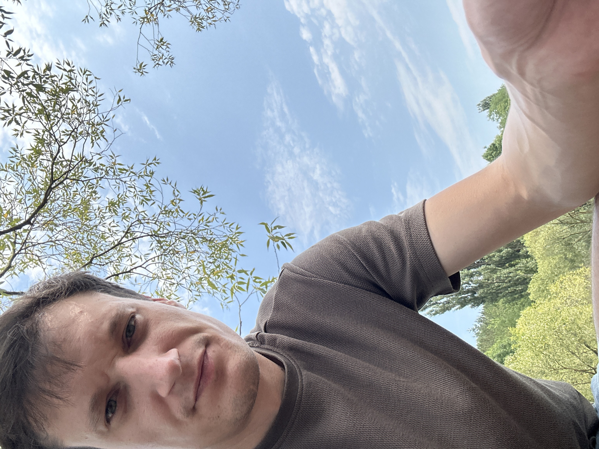 A man is taking a selfie outdoors, with his face and chest filling the left side of the frame. He is looking towards the camera with a neutral expression. He is wearing a brown t-shirt with a textured pattern. The background consists of a clear blue sky with scattered white clouds, framed by green tree branches and leaves. Below the sky, a lush green forest landscape is visible, suggesting an outdoor, natural setting possibly near Makiv, Ukraine, given the commonality of such scenery in rural and semi-rural Ukrainian locations. The perspective suggests the man is lying down or sitting, looking upwards at the sky.