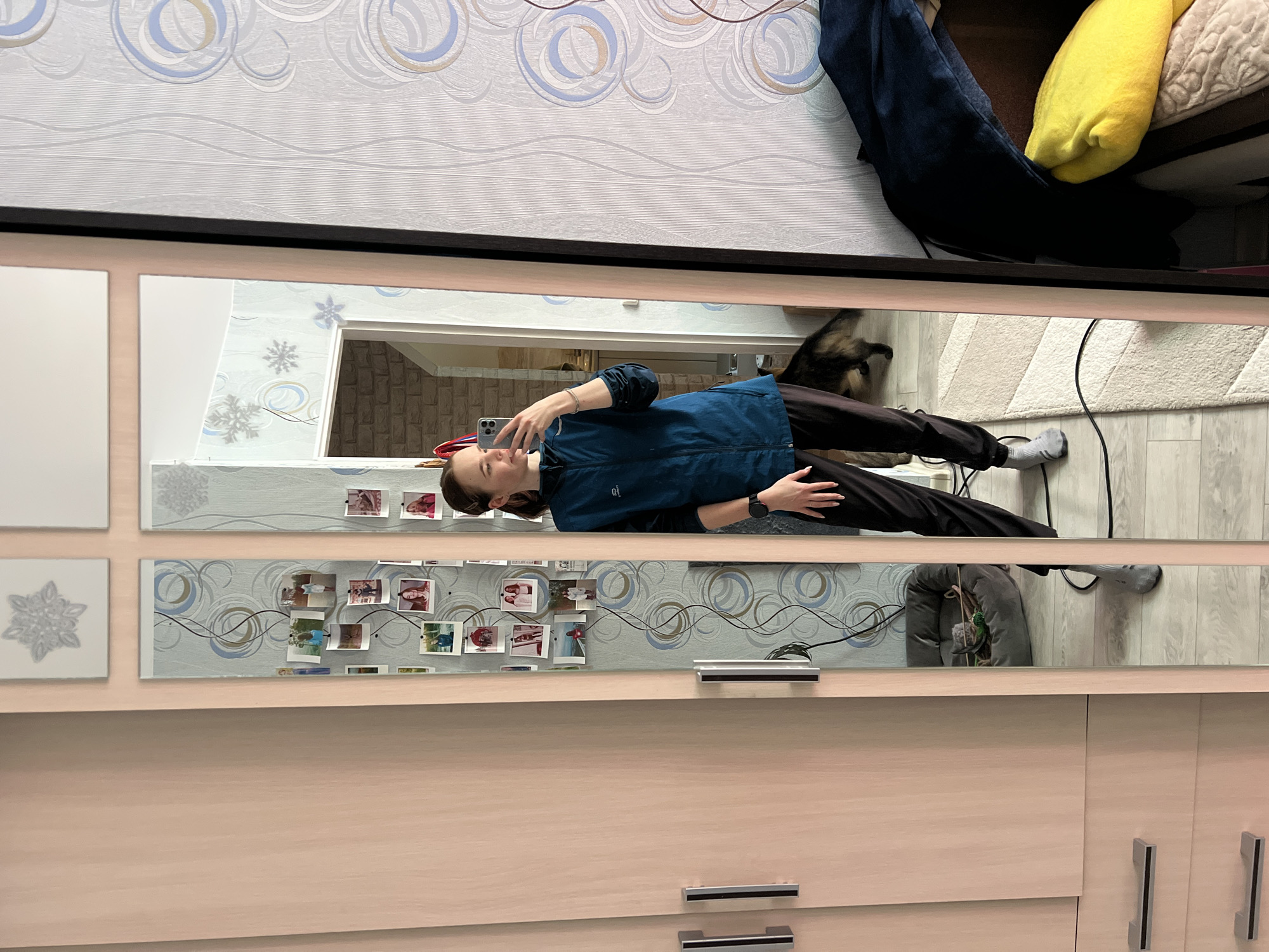 A mirrored reflection shows a woman lying on the floor, using a phone. She is wearing a dark blue jacket, black pants, and grey socks. The reflection also includes a dog, a grey chair, a white rug, and a cabinet with drawers. The wall above the mirror has wallpaper with a circular pattern. The reflection suggests the woman is in a room in Chaikovsky, Russia.