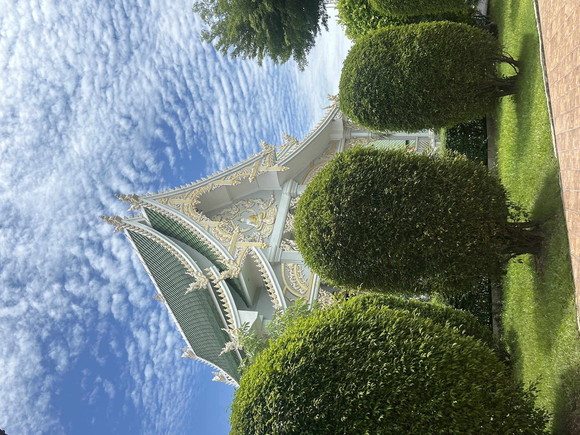 An exterior, eye-level, medium shot captures a Buddhist temple in Thai Ko Subdistrict, Thailand, under a bright, partly cloudy sky. The temple's ornate, white facade features intricate gold detailing and a vibrant green tiled roof. The building is partially obscured by lush, manicured green bushes in the foreground, which are shaped into rounded forms. The ground in front of the bushes is covered in green grass. To the right of the temple, a tall, leafy green tree stands. The overall scene is serene and depicts the architecture and landscaping of a religious site in a tropical climate.