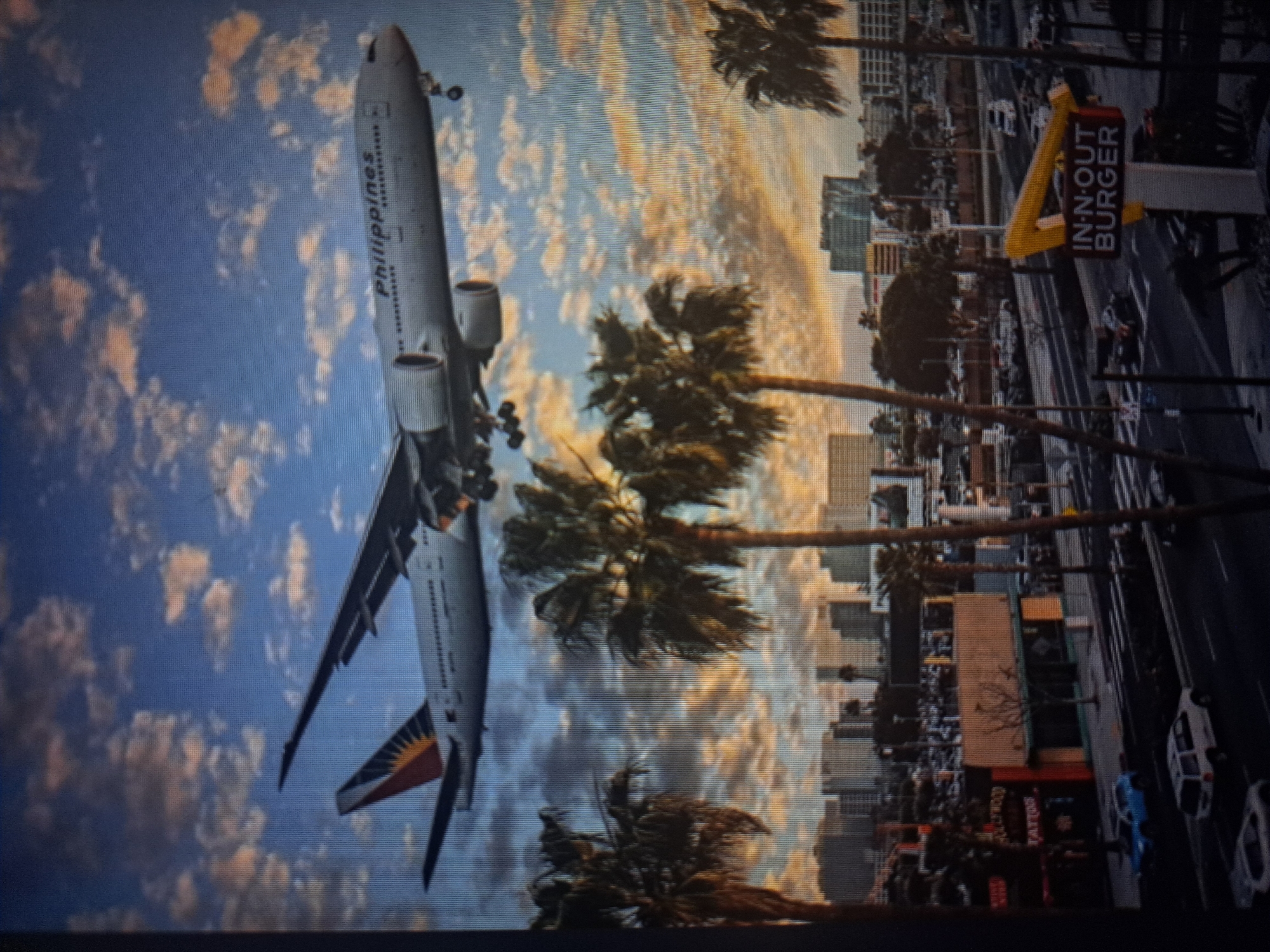 The image depicts a Philippines Airlines airplane approaching a city for landing, juxtaposed with an urban scene. The sky is filled with clouds, tinged with hues of orange and blue. Palm trees line the cityscape. In the foreground, an In-N-Out Burger sign is visible, suggesting a location in California, USA. The airplane is in landing configuration. The photograph is taken at an angle. There is no indication that the scene is in Yerevan, Armenia.