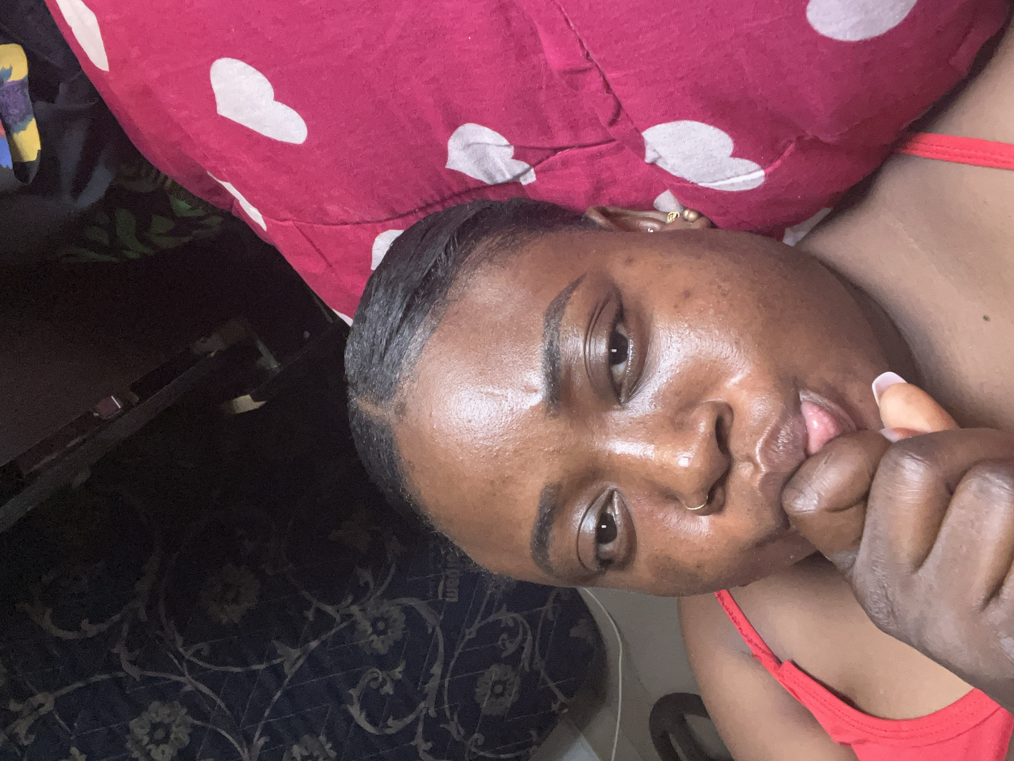 A close-up, slightly angled image shows a woman of color with dark hair pulled back from her face, lying on what appears to be a bed with a red pillow patterned with white hearts. She is wearing a red top. Her skin has a noticeable sheen. Her right hand is close to her face, with her fingers near her mouth. The background includes a dark area with a patterned rug or carpet. There is insufficient information to ascertain the location in Abuja, Nigeria.