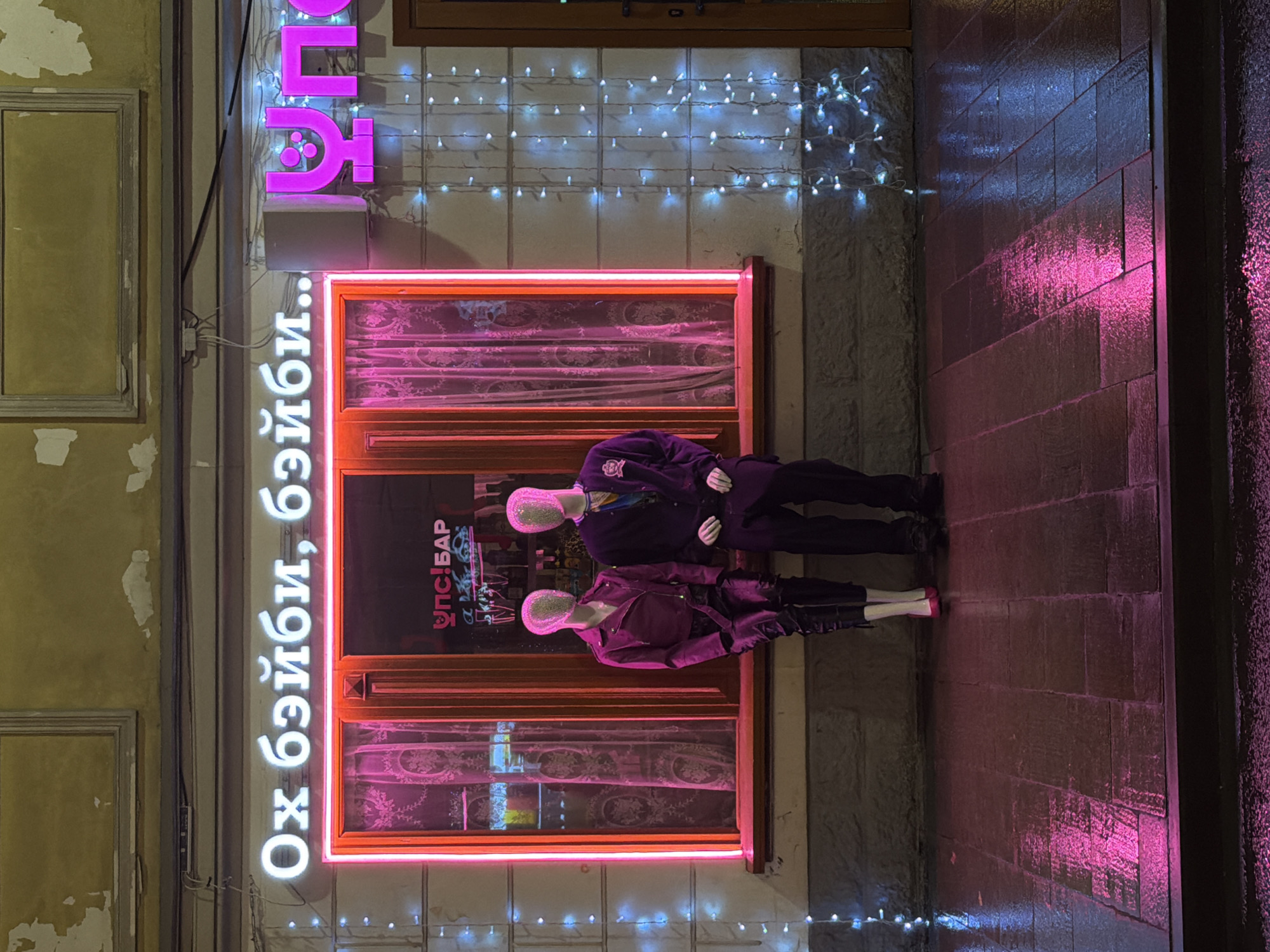 A street scene in Moscow, Russia, at night. The view is of a storefront featuring a neon sign in Russian that reads "Ox бэйби, бэйби.." vertically, and above the door, a neon pink sign with symbols. A neon-framed window display contains two mannequins dressed in dark clothing with glittering heads. One mannequin is wearing a dark suit with a police badge visible, and the other is wearing a jacket and pants. The window also contains a graphic that reads "Упс!Бар." Strings of small white lights are draped above the window. The wall to the left of the storefront appears worn, with peeling paint, and has rectangular frame-like indentations. The sidewalk in front of the store is made of dark, wet bricks that reflect the pink neon lights.
