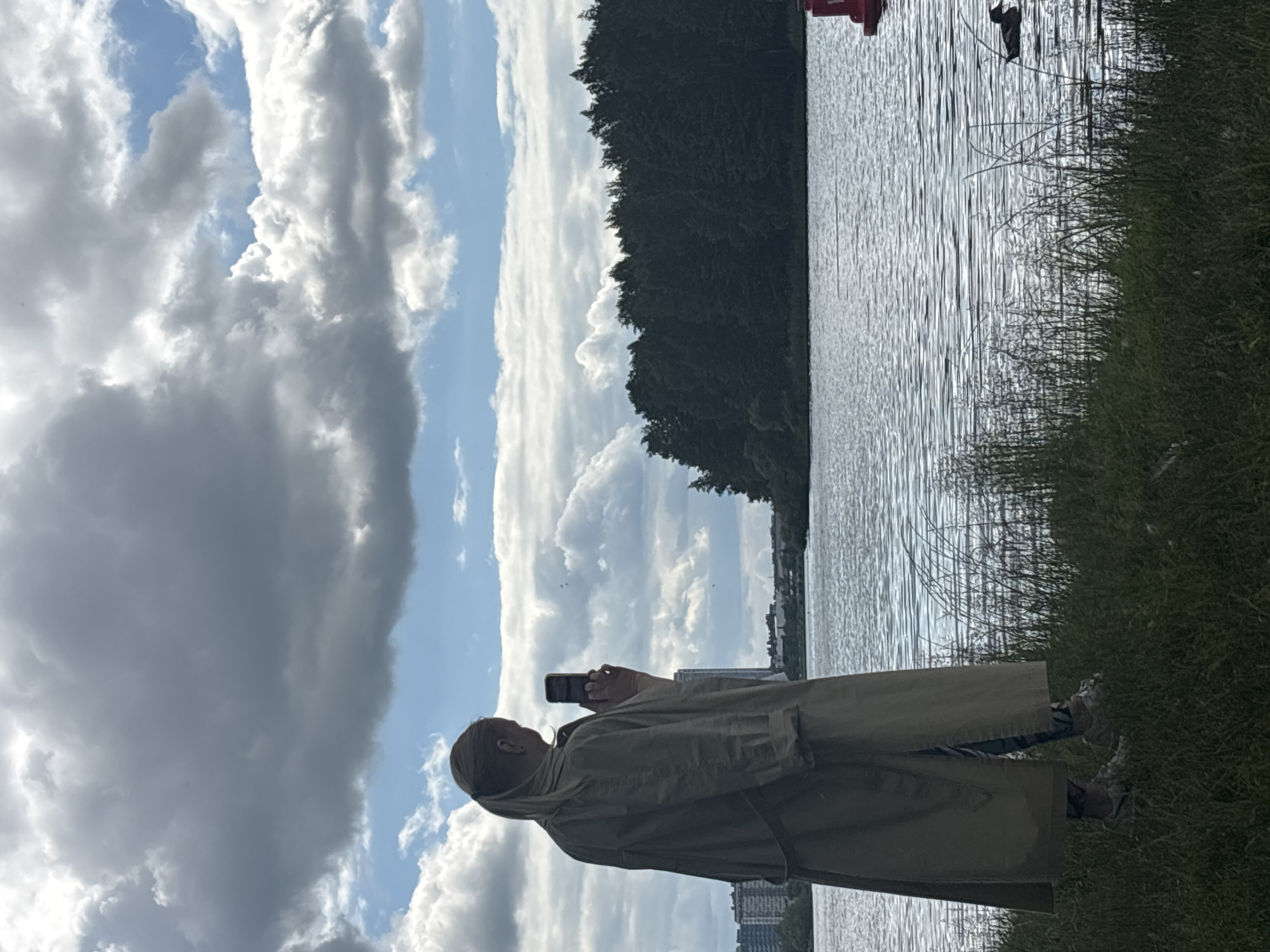 The image shows a person standing on the grassy bank of a body of water, holding a smartphone and appearing to take a photo or video. The person is wearing a light-colored trench coat. The water is rippling, reflecting the sky, and is bordered by a dense line of trees on the opposite bank. In the distance, several tall buildings are visible, suggesting an urban or suburban setting near a natural landscape. The sky is partially cloudy with patches of blue sky and dramatic, voluminous clouds. The overall impression is a moderately bright day with dynamic cloud formations. The scene suggests a moment of leisure or exploration, with the person capturing the scenery. The metadata indicates the location is Minsk, Belarus. There is no visible text in the image.