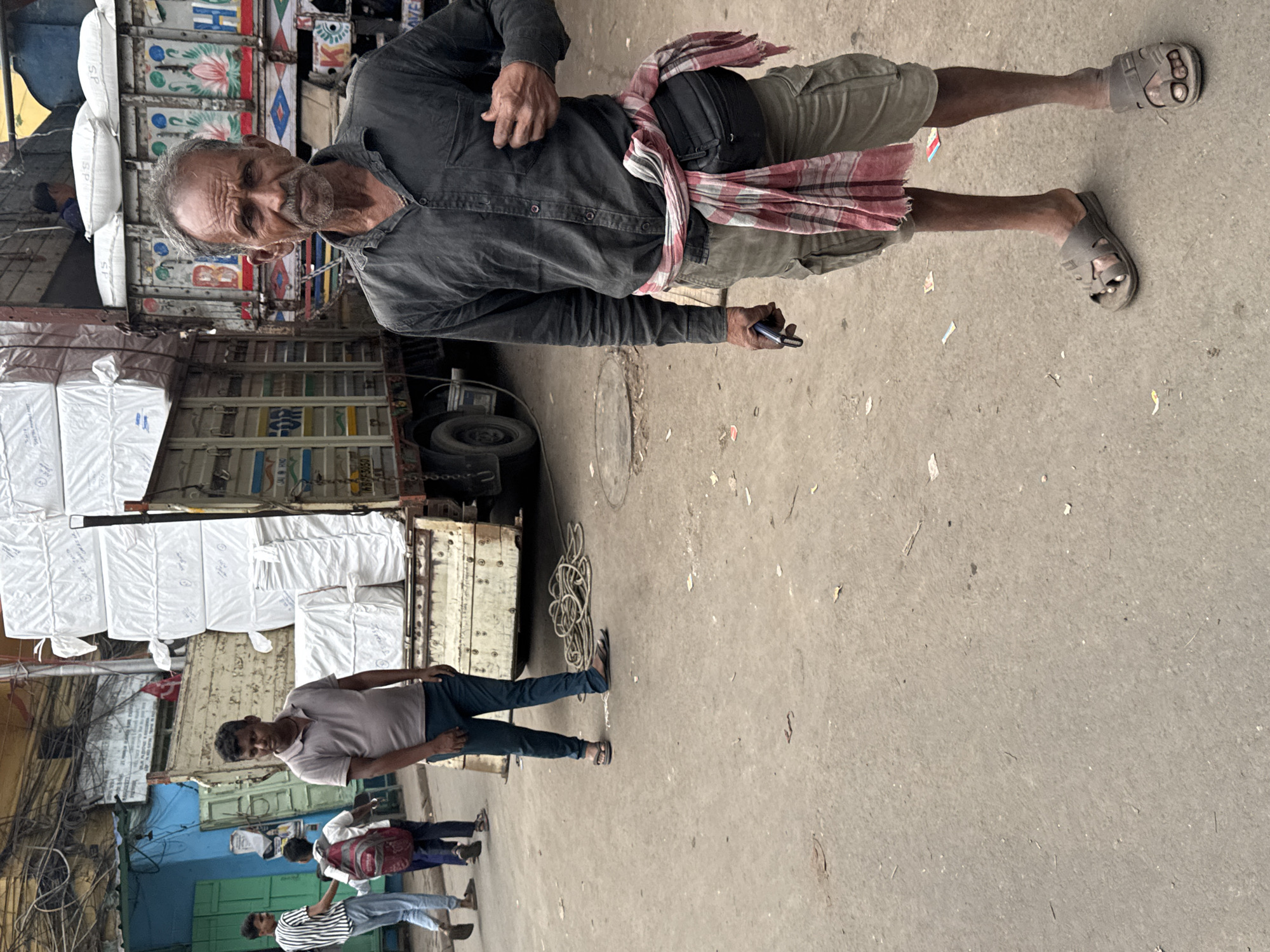 The image captures a scene in Kolkata, India. An elderly man, dressed in a dark shirt, light shorts, and sandals, stands near a truck loaded with bags. He holds a mobile phone. A red and white checkered cloth is tied around his waist. Several other people are seen walking on the street, including a man in a grey shirt and jeans. The background features buildings, a truck loaded with merchandise, and overhead power lines. The street is concrete with litter.