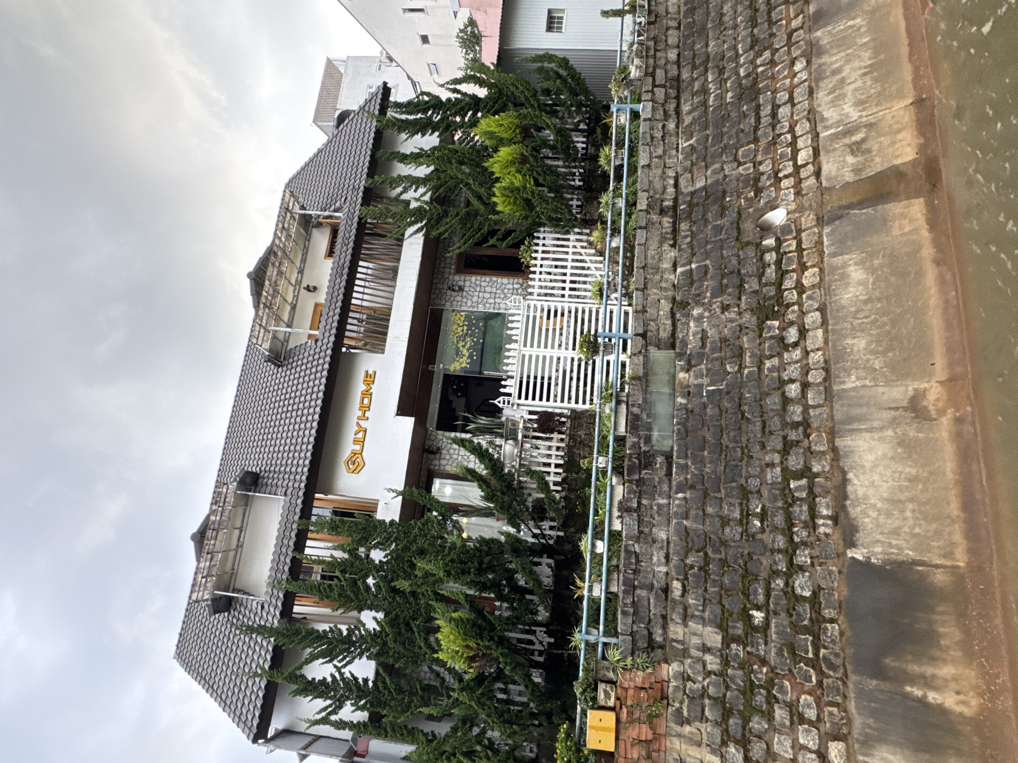 The image captures the exterior of a building named "Suly Home" in Da Lat, Vietnam. The building features a white facade with a dark tiled roof and a wooden balcony. Dense green coniferous trees flank the building's front, partially obscuring the entrance, which is bordered by a white picket fence. The structure is situated adjacent to a cobblestone path and a waterway, its edge visible on the right side of the frame. The weather appears overcast, as evidenced by the dull sky. No people are visible in the scene.
