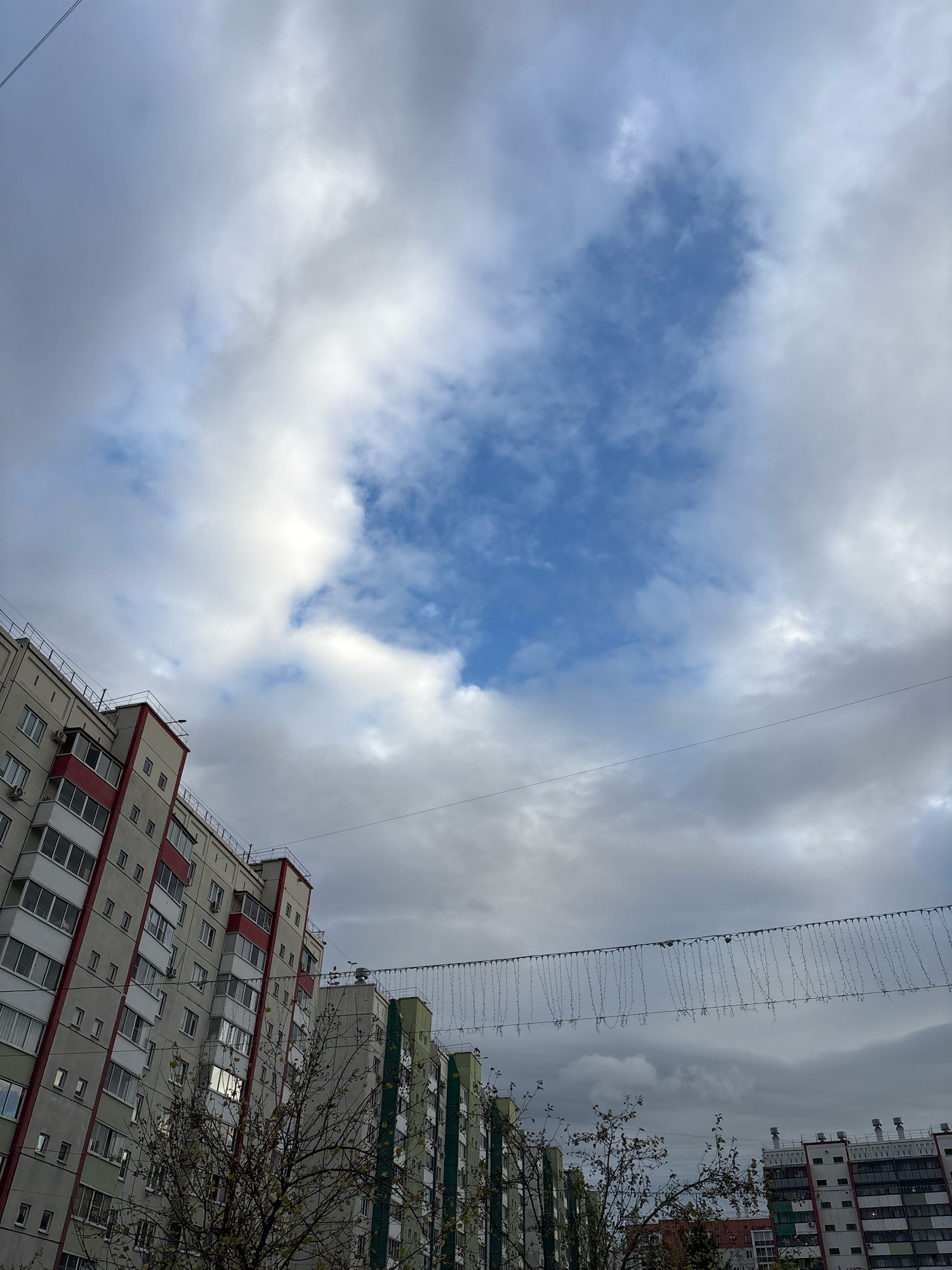 The image depicts an overcast sky above residential buildings in the city of Zapadny, Russia. The buildings are multi-story apartment blocks with visible windows and balconies. Some have a light gray facade with red and green accents, while others are primarily green. A line of string lights, likely decorative, is suspended across the frame between two buildings. Bare tree branches are visible in the foreground. The overall atmosphere is somber, with the heavy cloud cover dominating the sky. No people or specific actions are discernible.