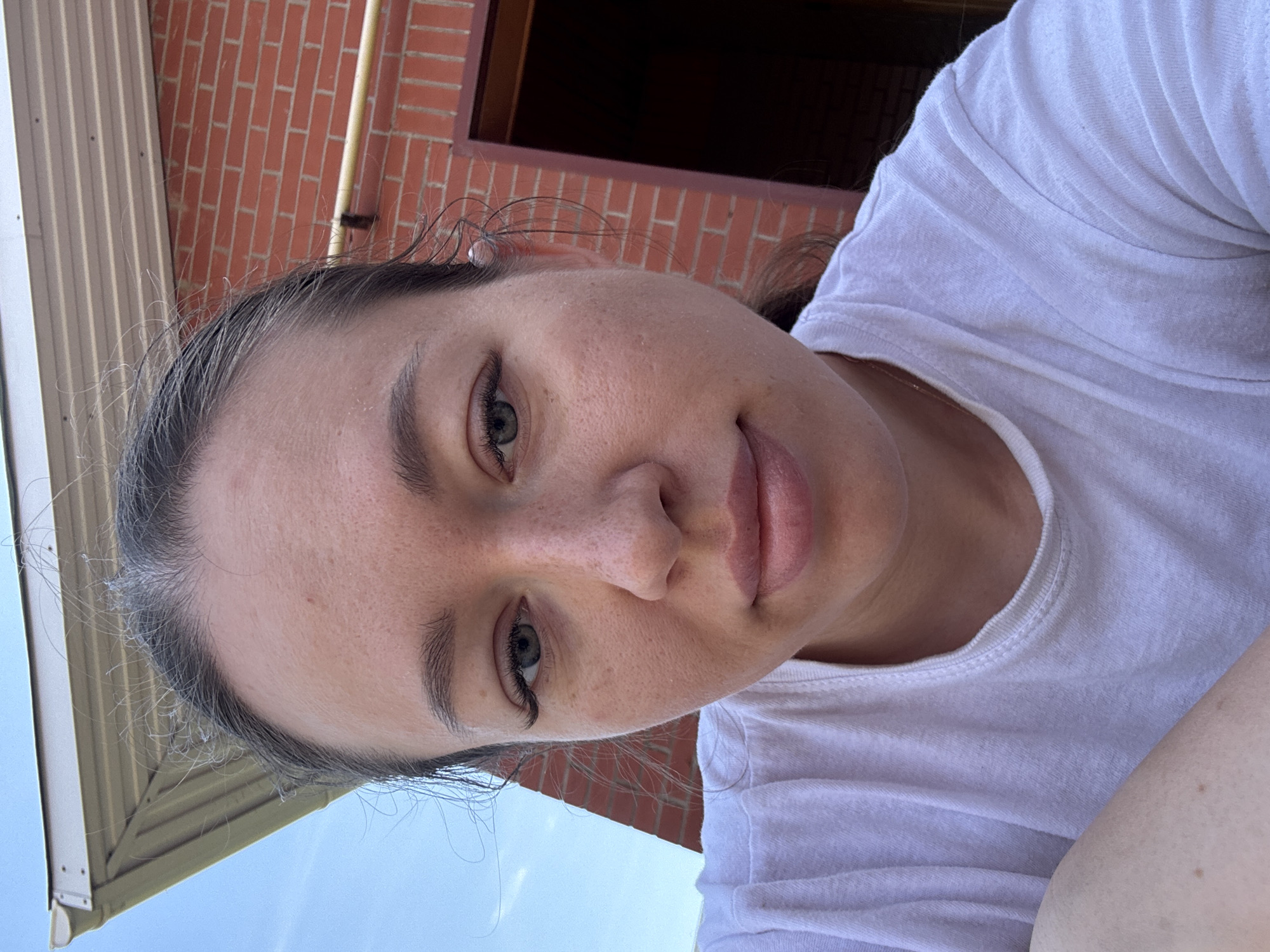 The image shows a woman with fair skin and graying dark hair wearing a white t-shirt. She is looking at the camera with a neutral expression. The background includes a brick building and a blue sky. Based on the information provided, the location is in Ферма, Russia.