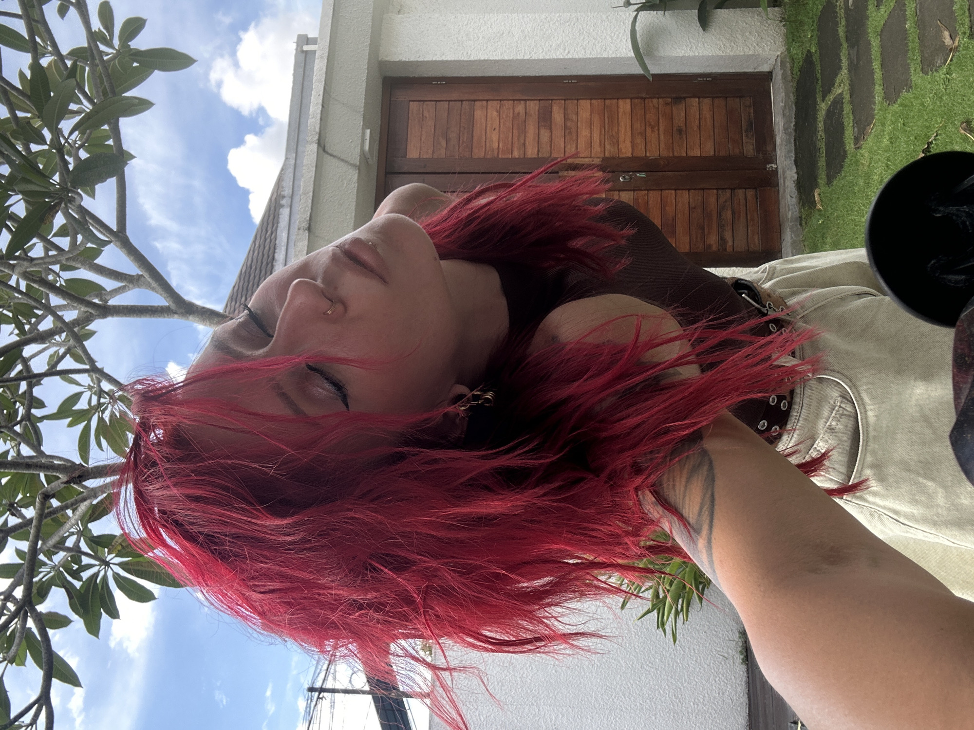 The image displays a person with vibrant, long red hair, captured in a low-angle shot. Their head is tilted back, and their eyes are closed, conveying a sense of relaxation or introspection. A small nose ring and a gold hoop earring are visible. A partial view of a dark-lined tattoo adorns their left arm, and they wear a light-colored, possibly beige or khaki, garment with a dark strap across the chest.
The background reveals a bright blue sky with scattered white clouds, indicating a clear, sunny day. To the left, branches of a tree with large green leaves, likely tropical foliage such as frangipani, are prominent. On the right, a segment of a white building is visible, featuring a prominent wooden door or gate made of vertical planks. Below the building, a patch of green grass with irregular stone pavers suggests a garden path. A dark, circular object is partially visible in the lower right corner of the frame.
The setting appears to be outdoors in a tropical environment, consistent with the specified location of North Kuta, Indonesia. The overall mood is peaceful and serene. No visible text is present in the image.