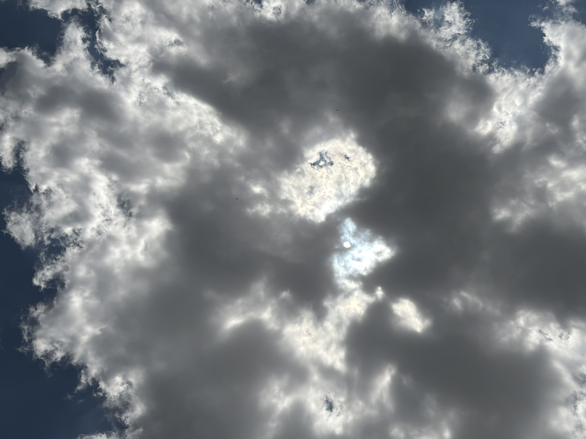 A low-angle shot looks up at a sky filled with dark, voluminous clouds. The sun shines through a break in the clouds, creating a bright, hazy glow. The sunlight illuminates the edges of the clouds, highlighting their textures and contours. The sky is a deep blue around the clouds. No people, subjects, or objects are visible. The location is Oxentea, Moldova.