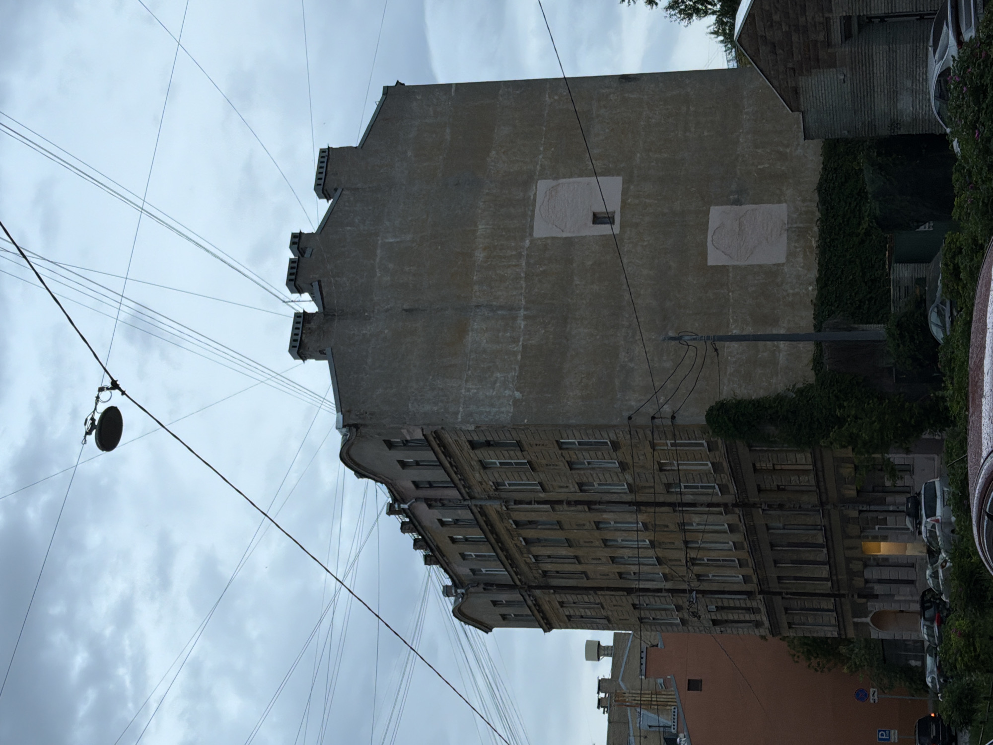 The image depicts a cloudy day in Saint Petersburg, Russia, with a tall, multi-story building dominating the frame. The building appears to be made of aged brick and plaster, with numerous windows arranged in a grid pattern. Several electrical wires crisscross the sky, strung between poles and buildings. A street lamp is visible hanging from one of these wires. Two light-colored, irregularly shaped patches are present on the large, blank side of the building, one of which has a small, dark square visible within it, possibly a tiny window. The lower section of the building is partially obscured by dense green foliage and trees. In the foreground, several cars are parked along a street, and more buildings, including one with a stone facade, can be seen to the right and left. A parking sign is visible on the left side of the image. The overall scene suggests an urban environment with older architecture, typical of some districts in Saint Petersburg.