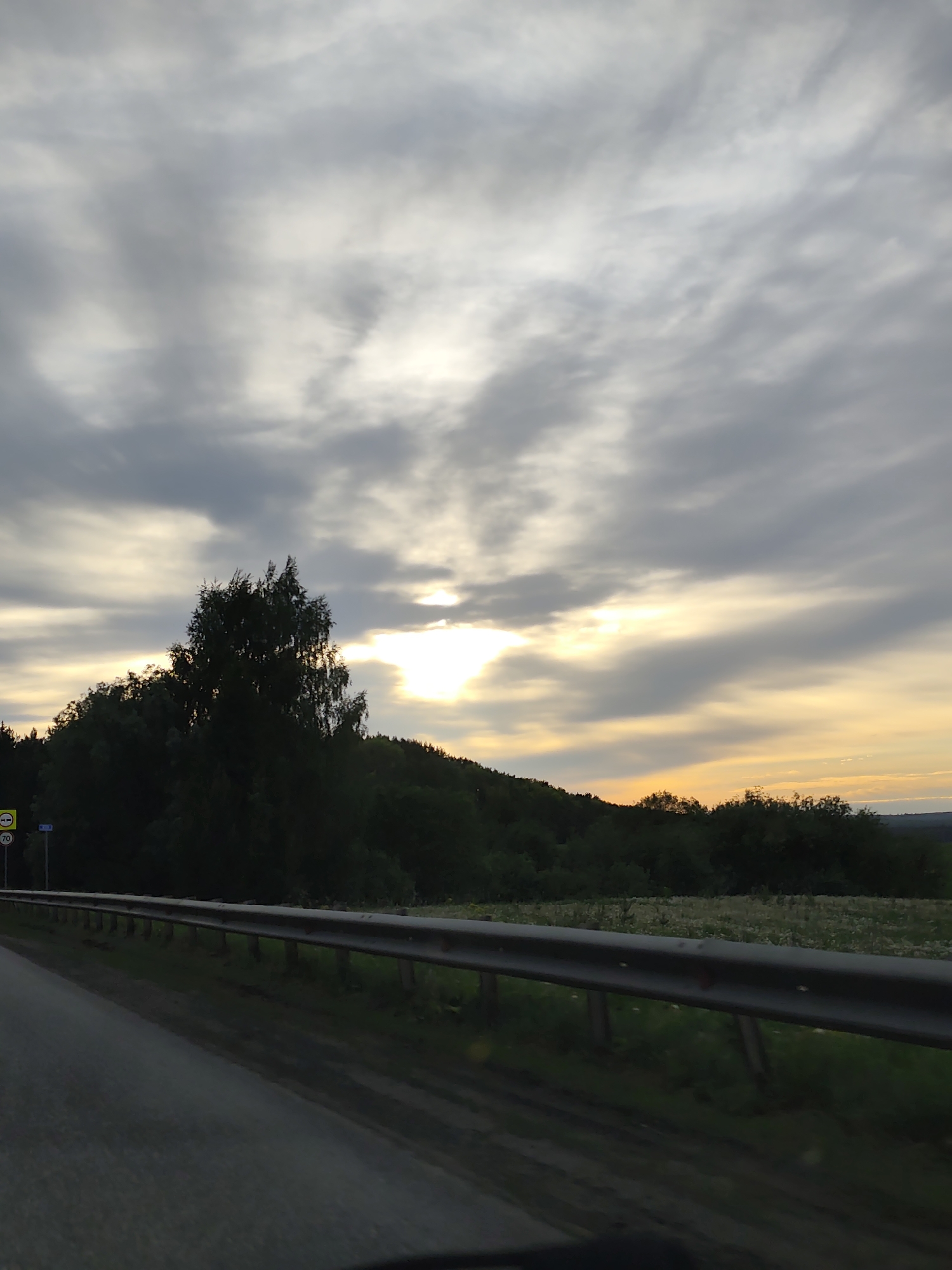 The image depicts a roadside view at dusk in the vicinity of Lukintsy, Russia. A two-lane road is visible in the foreground, separated from a grassy area by a metal guardrail. Beyond the guardrail, there is a field with wildflowers, backed by a densely forested hill. The sky is overcast with stratus clouds, and the setting sun peeks through a break in the clouds, casting a warm glow along the horizon. Road signs with speed limits (70) can be seen.